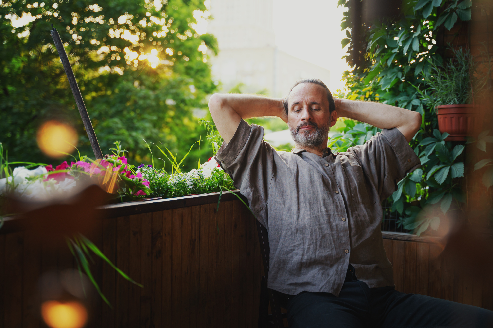 A man with a relaxed expression sits on a wooden balcony with his hands behind his head. He is wearing a loose, grey shirt. The balcony is adorned with plants, flowers, and greenery, and the background shows trees illuminated by sunlight.