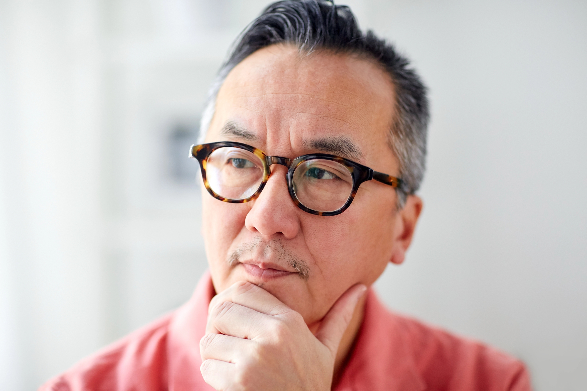 A man with short, graying hair and glasses is thoughtfully gazing into the distance. He rests his chin on his hand, wearing a coral shirt. The background is softly blurred, suggesting an indoor setting.