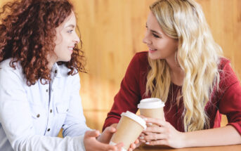 Two women are sitting at a table, smiling and holding takeaway coffee cups. One woman has curly red hair and is wearing a light blue shirt, the other has long blonde hair and is wearing a red blouse. They appear to be engaged in a friendly conversation.