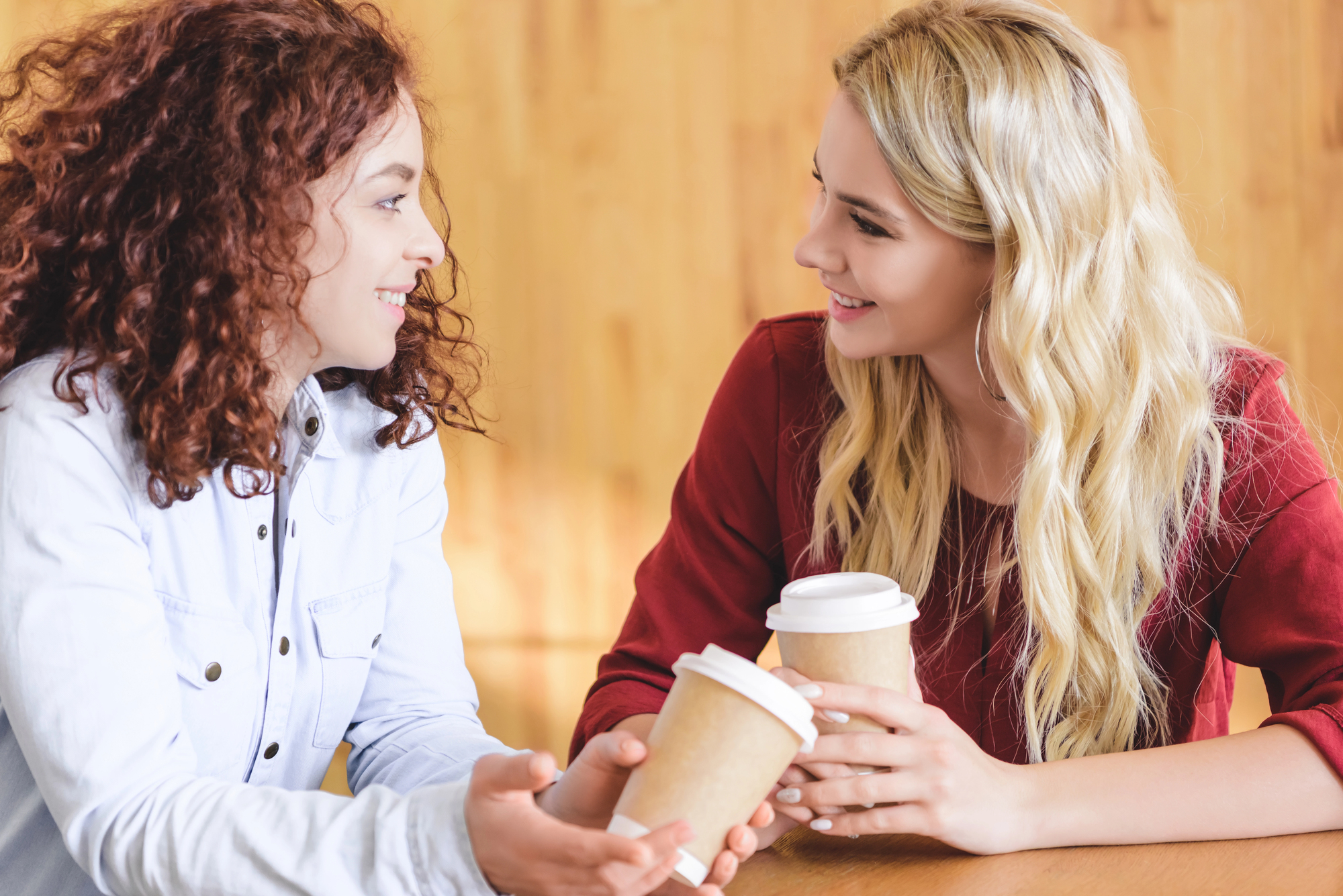 Two women are sitting at a table, smiling and holding takeaway coffee cups. One woman has curly red hair and is wearing a light blue shirt, the other has long blonde hair and is wearing a red blouse. They appear to be engaged in a friendly conversation.