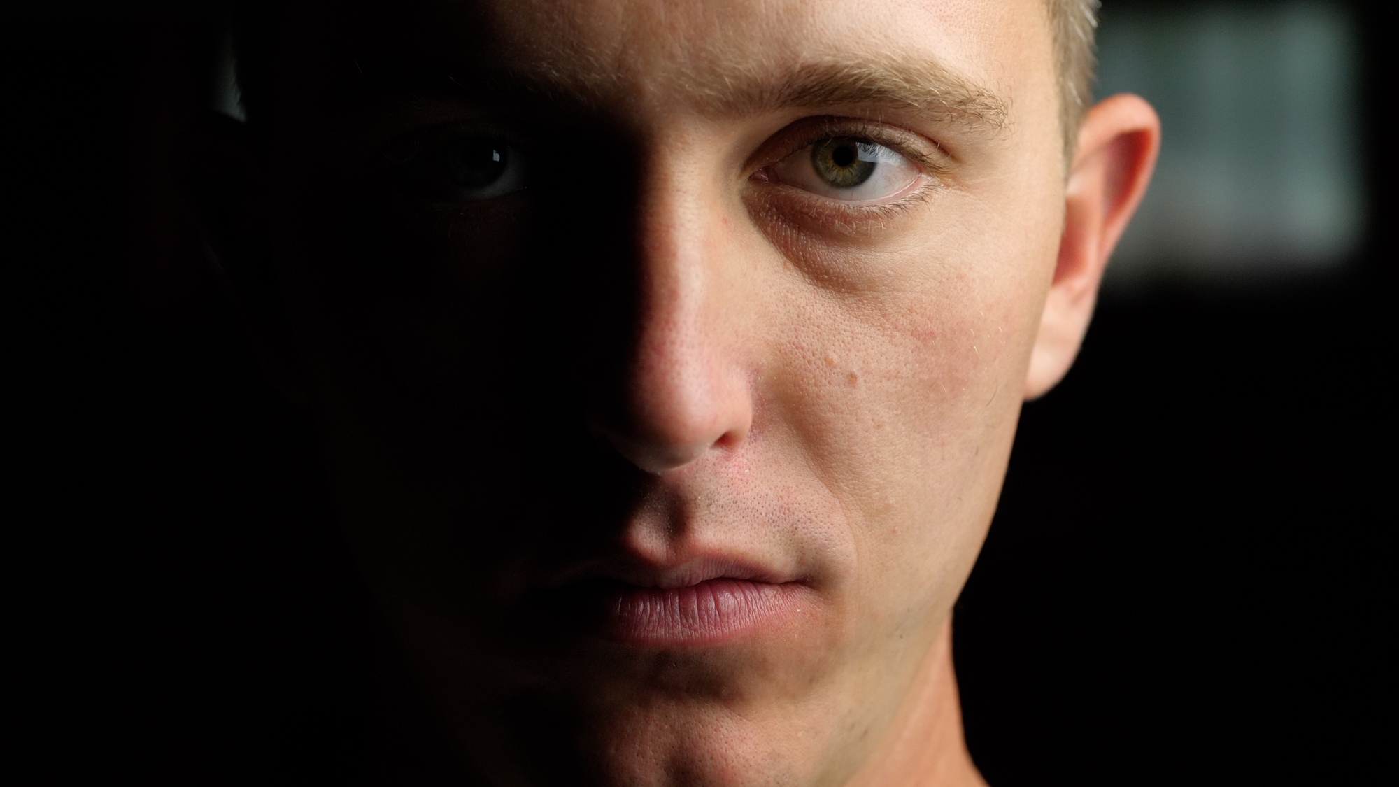 Close-up of a person with a neutral expression, facing the camera. The image is dimly lit with shadows on the left side of the person's face, highlighting their facial features and green eyes. The background is dark and indistinct.