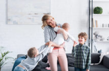 A woman smiles while holding a baby. She is standing in a bright living room. Two children, one gripping her arm and another standing nearby, interact playfully with her. The room has white walls, a gray couch, and plants and books on shelves in the background.