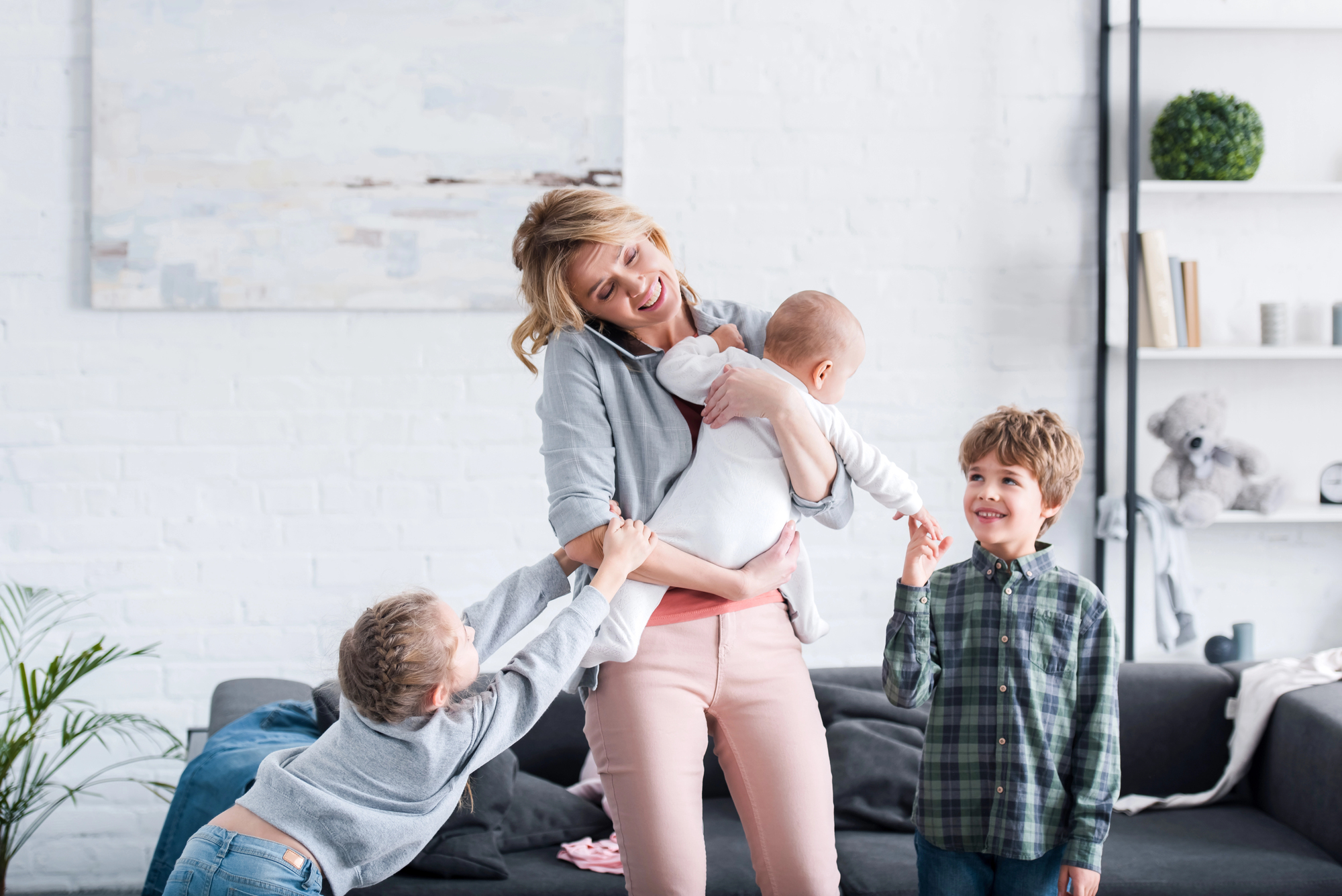 A woman smiles while holding a baby. She is standing in a bright living room. Two children, one gripping her arm and another standing nearby, interact playfully with her. The room has white walls, a gray couch, and plants and books on shelves in the background.