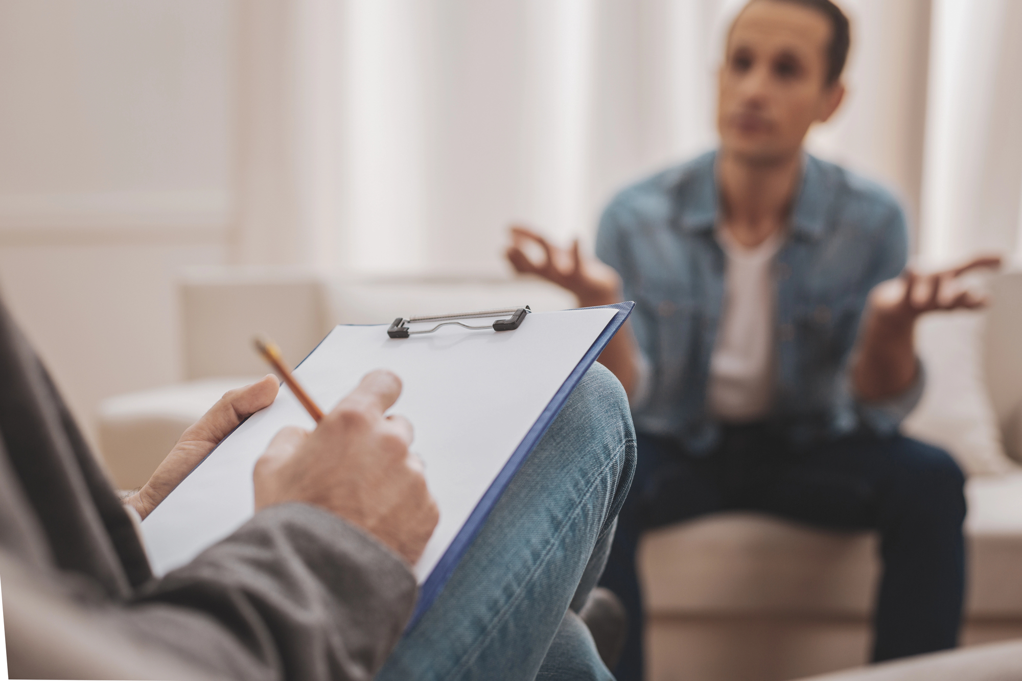 A close-up of a clipboard and pen held by an individual taking notes, with a blurred person in the background gesturing with both hands during what appears to be a conversation or consultation. The focus is on the note-taking activity in the foreground.