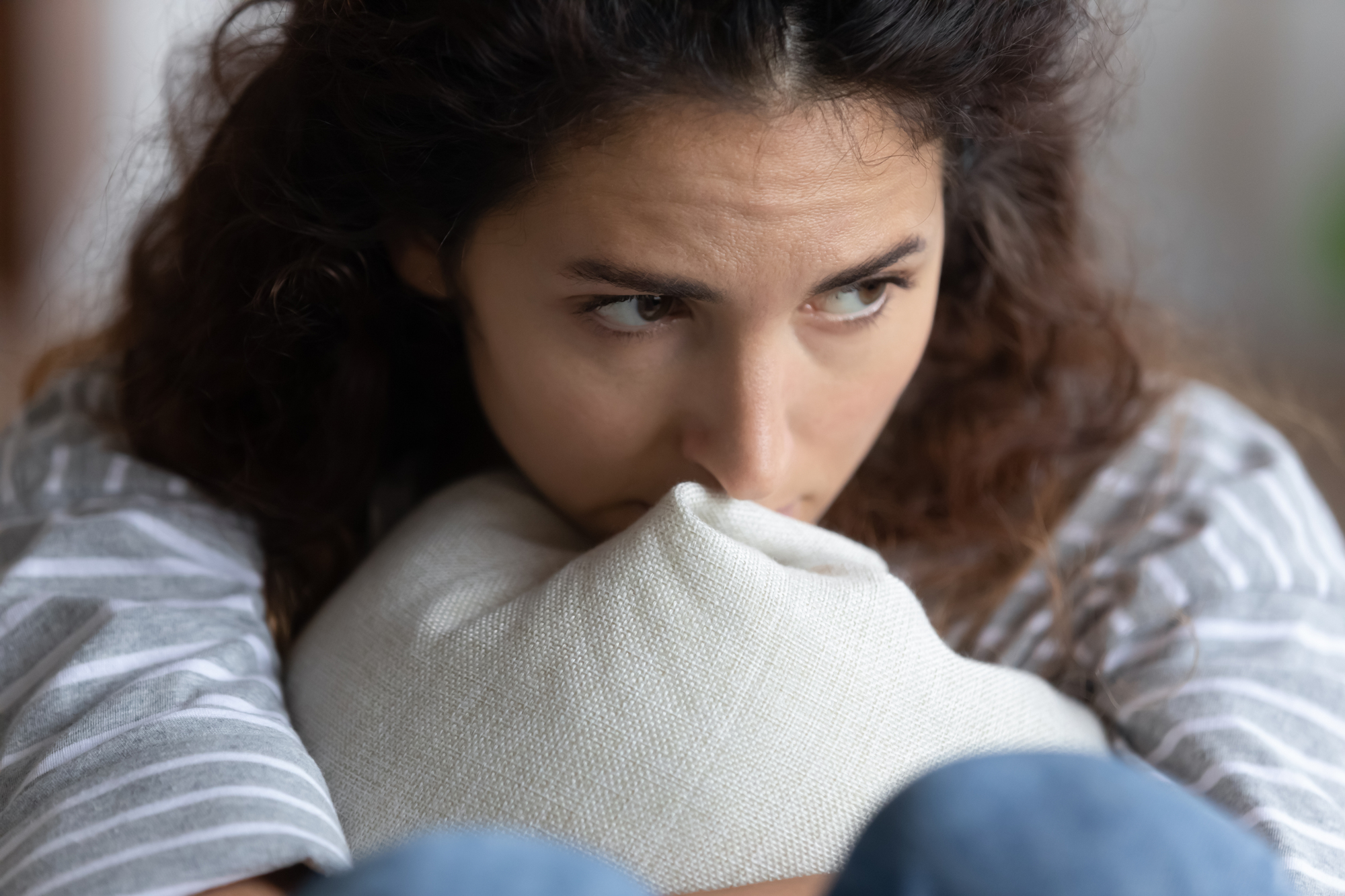 A woman with curly hair, wearing a striped top, sits with her knees up, clutching a light-colored cushion to her chest. She looks to the side with a pensive expression, appearing deep in thought.