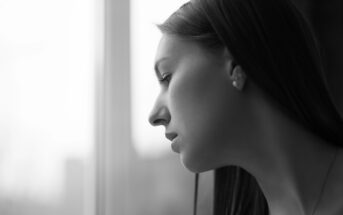 Black and white close-up of a woman with long hair looking out of a window. Her face is in profile, appearing thoughtful or contemplative. The background is blurred, giving a soft-focus effect to the overall image.