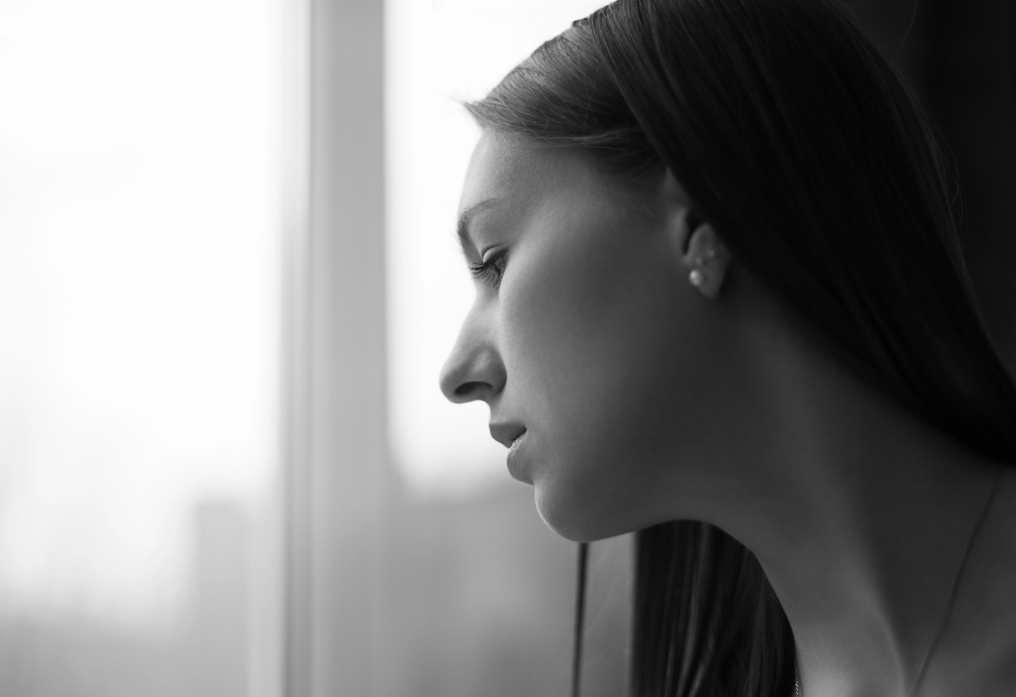 Black and white close-up of a woman with long hair looking out of a window. Her face is in profile, appearing thoughtful or contemplative. The background is blurred, giving a soft-focus effect to the overall image.