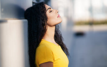 A woman with long curly hair is leaning against a wall with her eyes closed, head tilted back, and wearing a bright yellow shirt. The background is blurred, focusing attention on her relaxed and meditative expression.