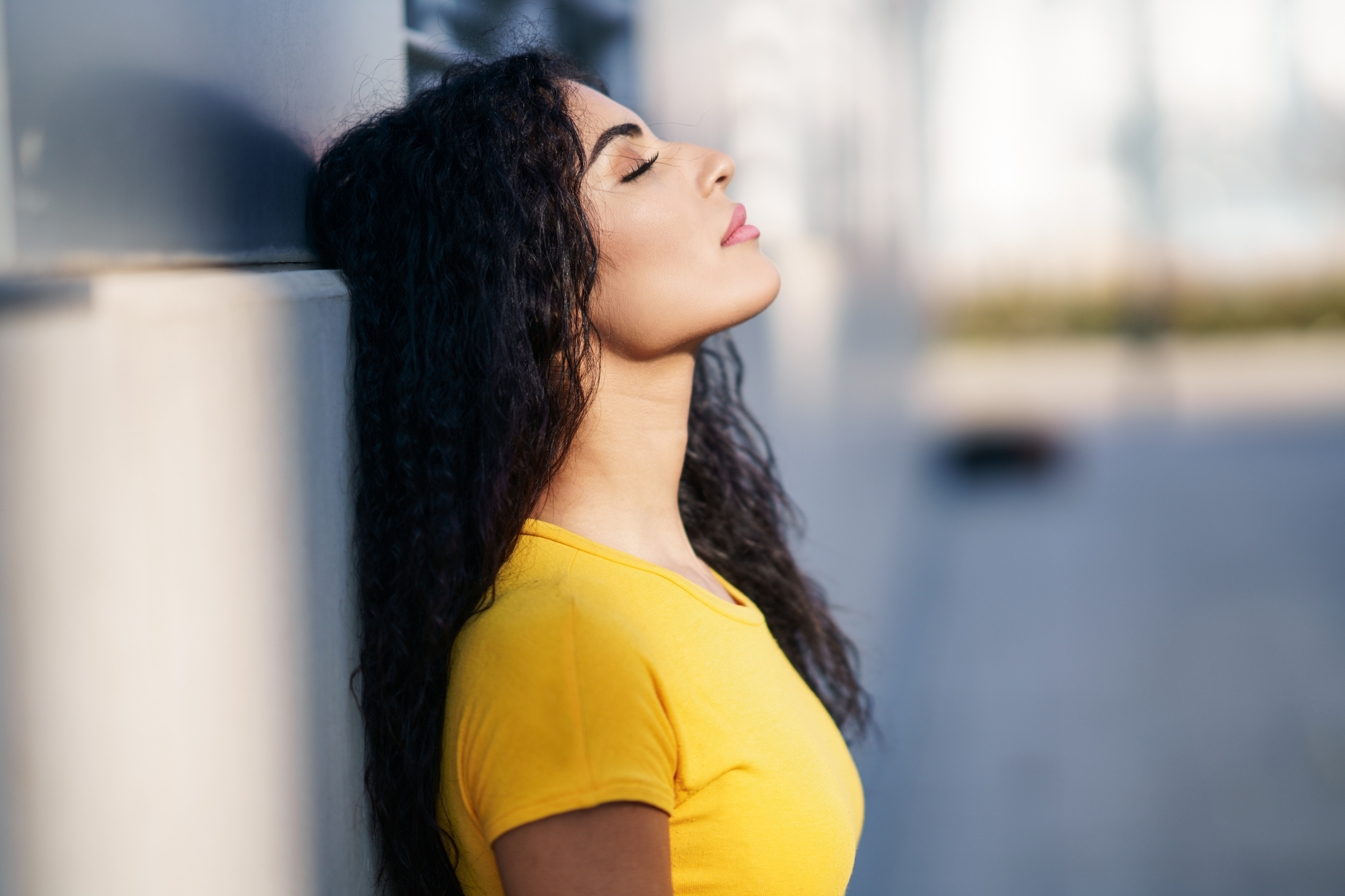 A woman with long curly hair is leaning against a wall with her eyes closed, head tilted back, and wearing a bright yellow shirt. The background is blurred, focusing attention on her relaxed and meditative expression.