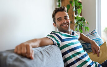 A man with short brown hair and a beard is sitting on a grey couch, smiling. He is wearing a striped shirt with blue, green, and white horizontal lines. There are cushions next to him, and potted plants can be seen in the background.