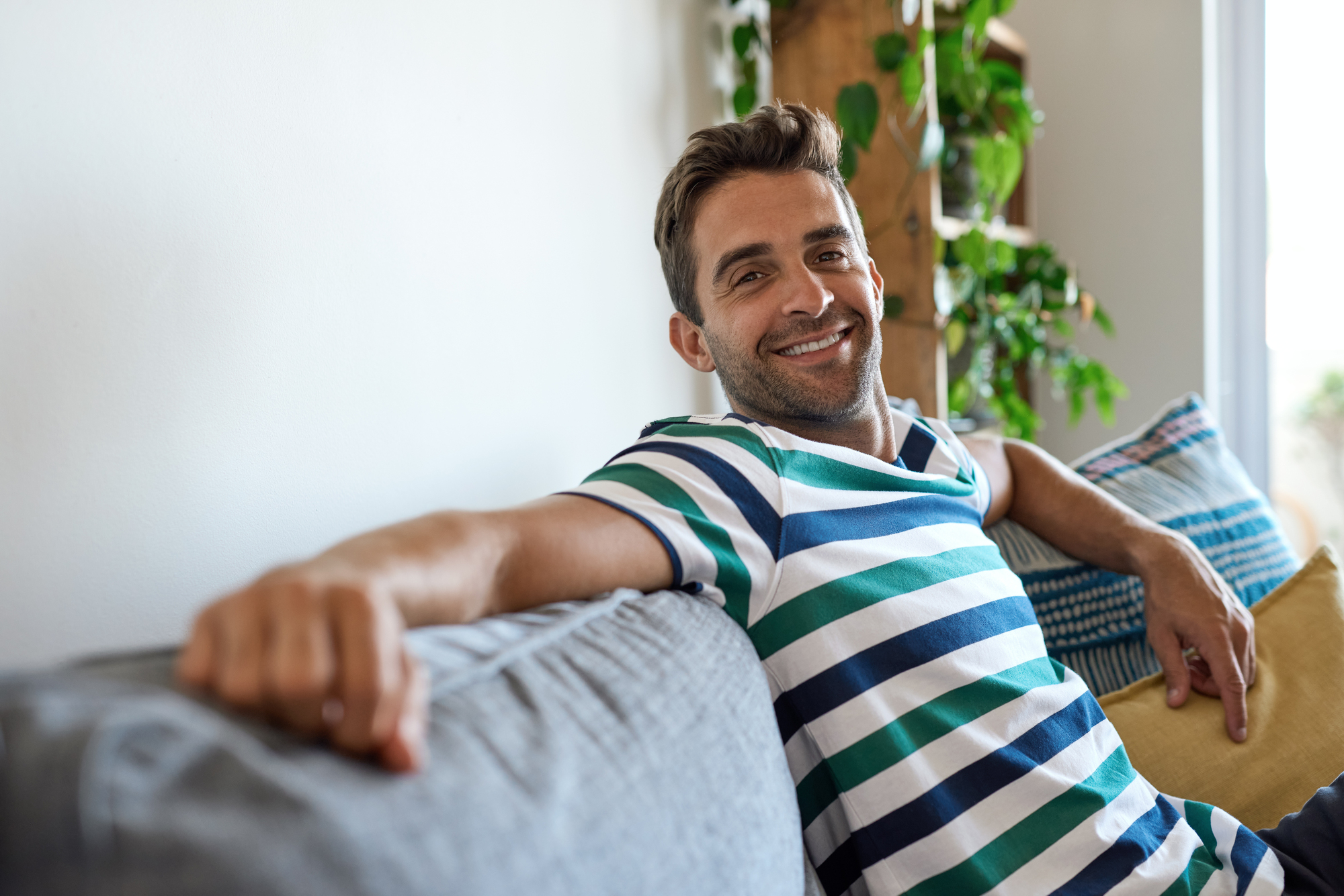 A man with short brown hair and a beard is sitting on a grey couch, smiling. He is wearing a striped shirt with blue, green, and white horizontal lines. There are cushions next to him, and potted plants can be seen in the background.