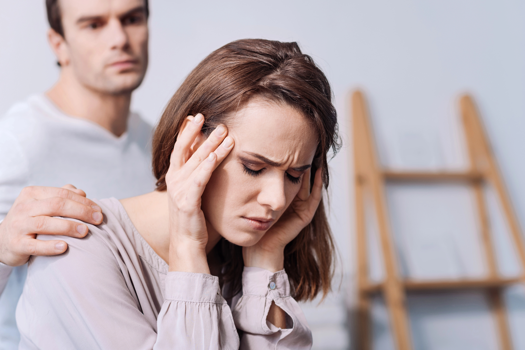 A woman with brown hair sits with her eyes closed, holding her temples with a pained expression. A man stands behind her with his hand on her shoulder, looking concerned. There is a blurred wooden easel in the background.