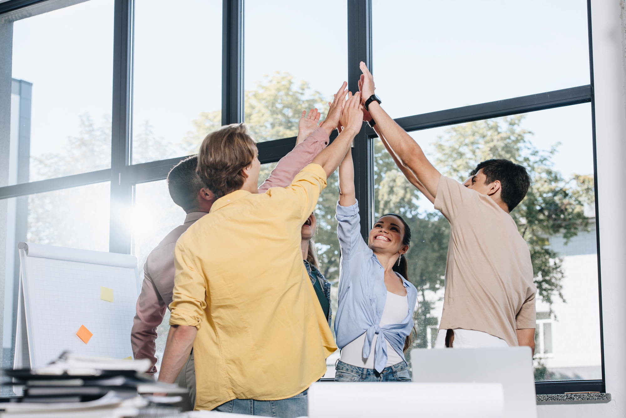 A group of five people stands in a circle inside a bright room with large windows. They are all smiling and giving a high-five simultaneously, celebrating something. There is a flipchart with sticky notes and a desk with papers in the foreground.