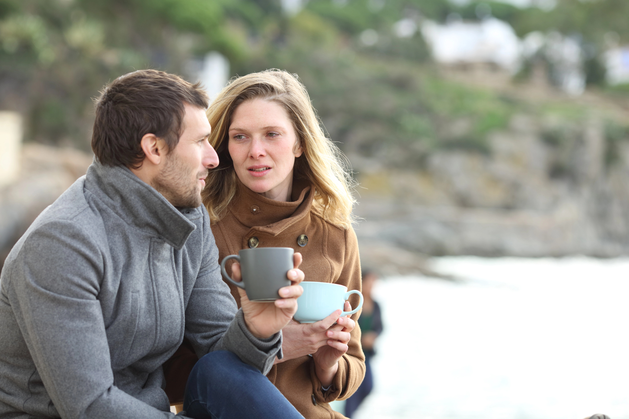 A man and a woman are sitting outdoors, wearing coats and holding mugs, seemingly engaged in a conversation. Behind them is a blurred natural background with greenery and water. The woman is looking at the man while he speaks.