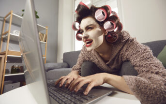 A person with facial cream and hair curlers types on a laptop while sitting on a couch. They are wearing a cozy robe and have an intense, focused expression. The background includes shelves with books and decorative items.