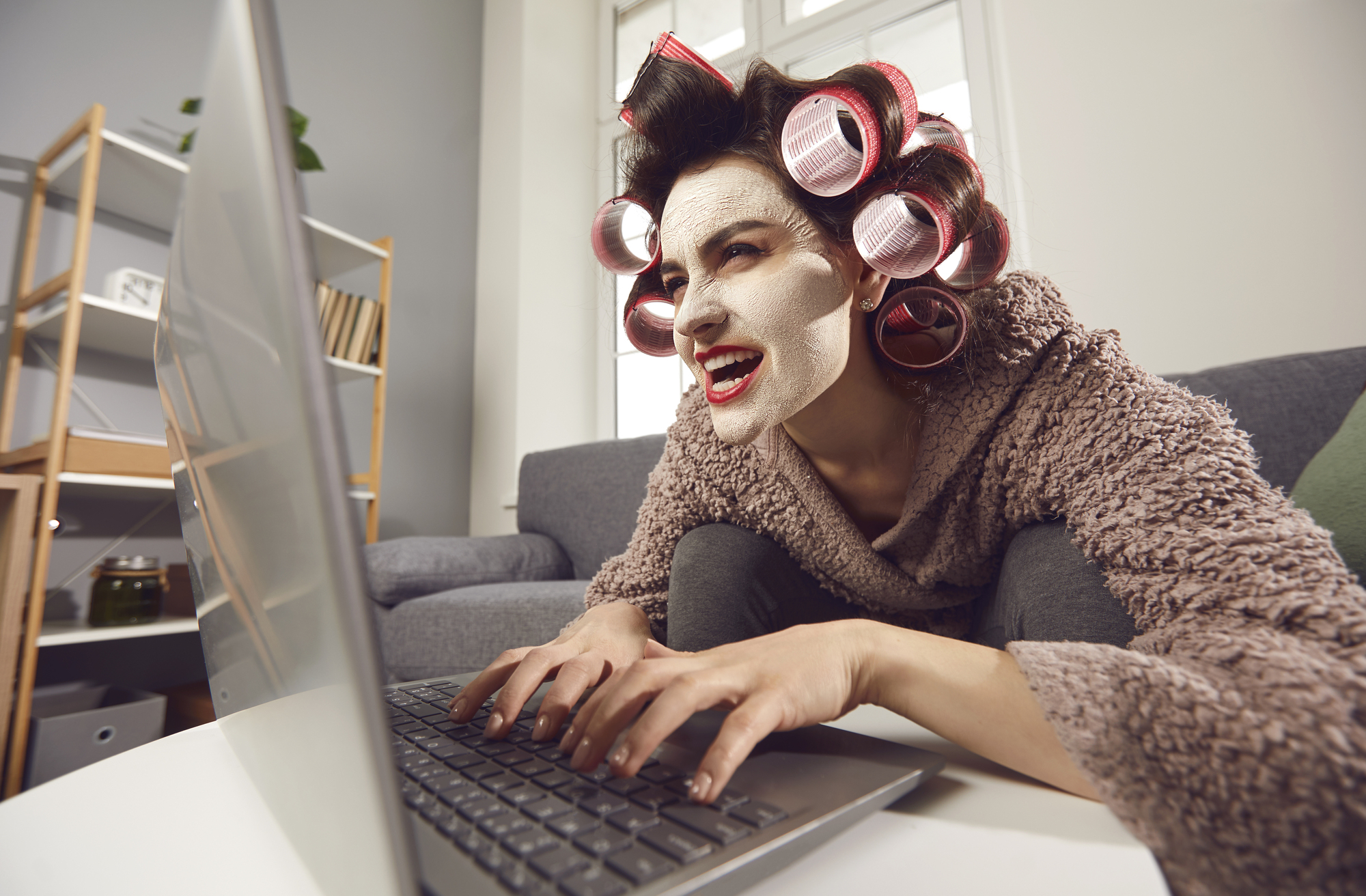 A person with facial cream and hair curlers types on a laptop while sitting on a couch. They are wearing a cozy robe and have an intense, focused expression. The background includes shelves with books and decorative items.