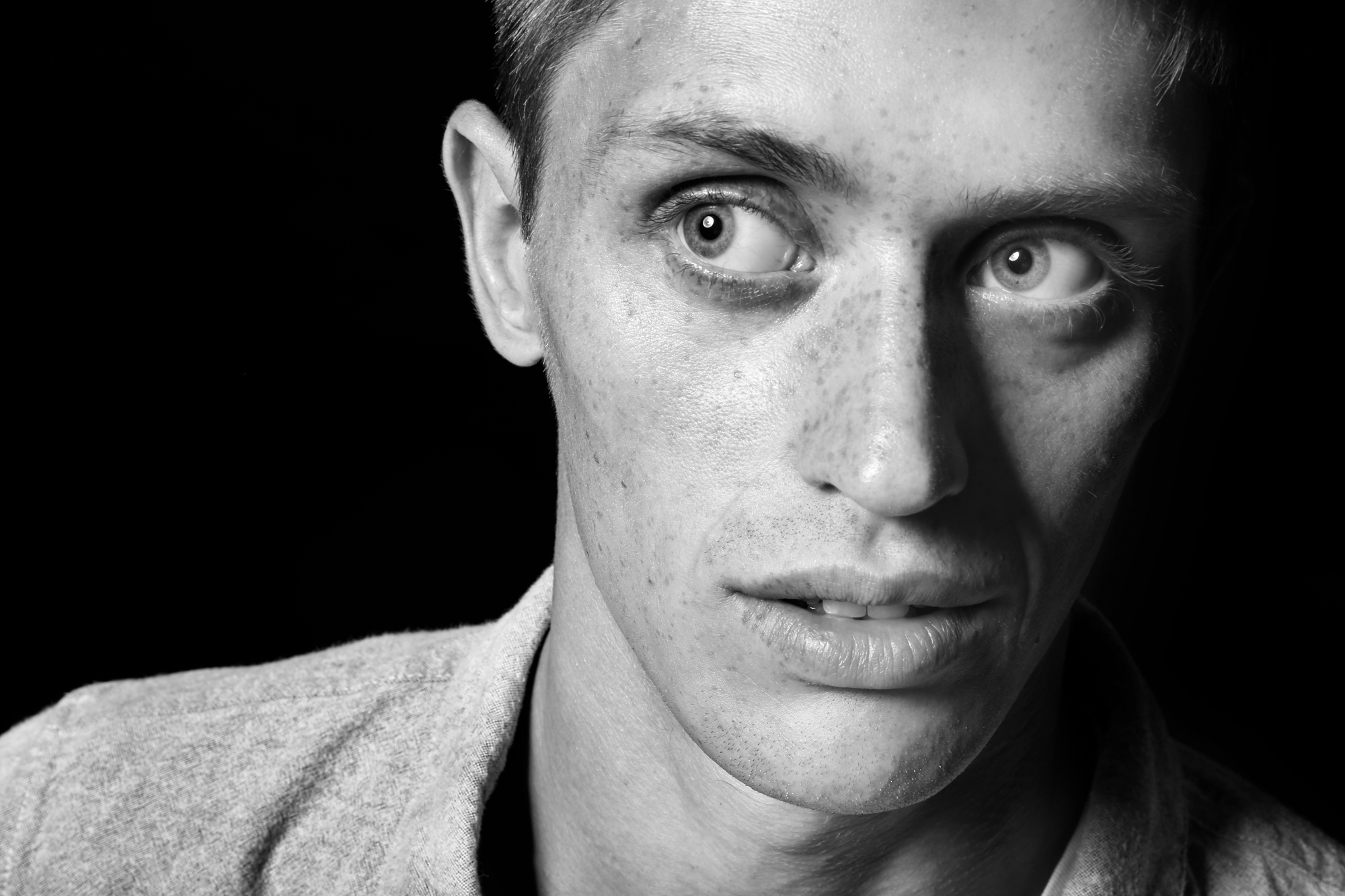 A black and white close-up portrait of a person looking slightly to the right. The face is partially shadowed, highlighting the person's intense, wide-eyed expression. They have short hair and are wearing a light-colored collared shirt. The background is black.