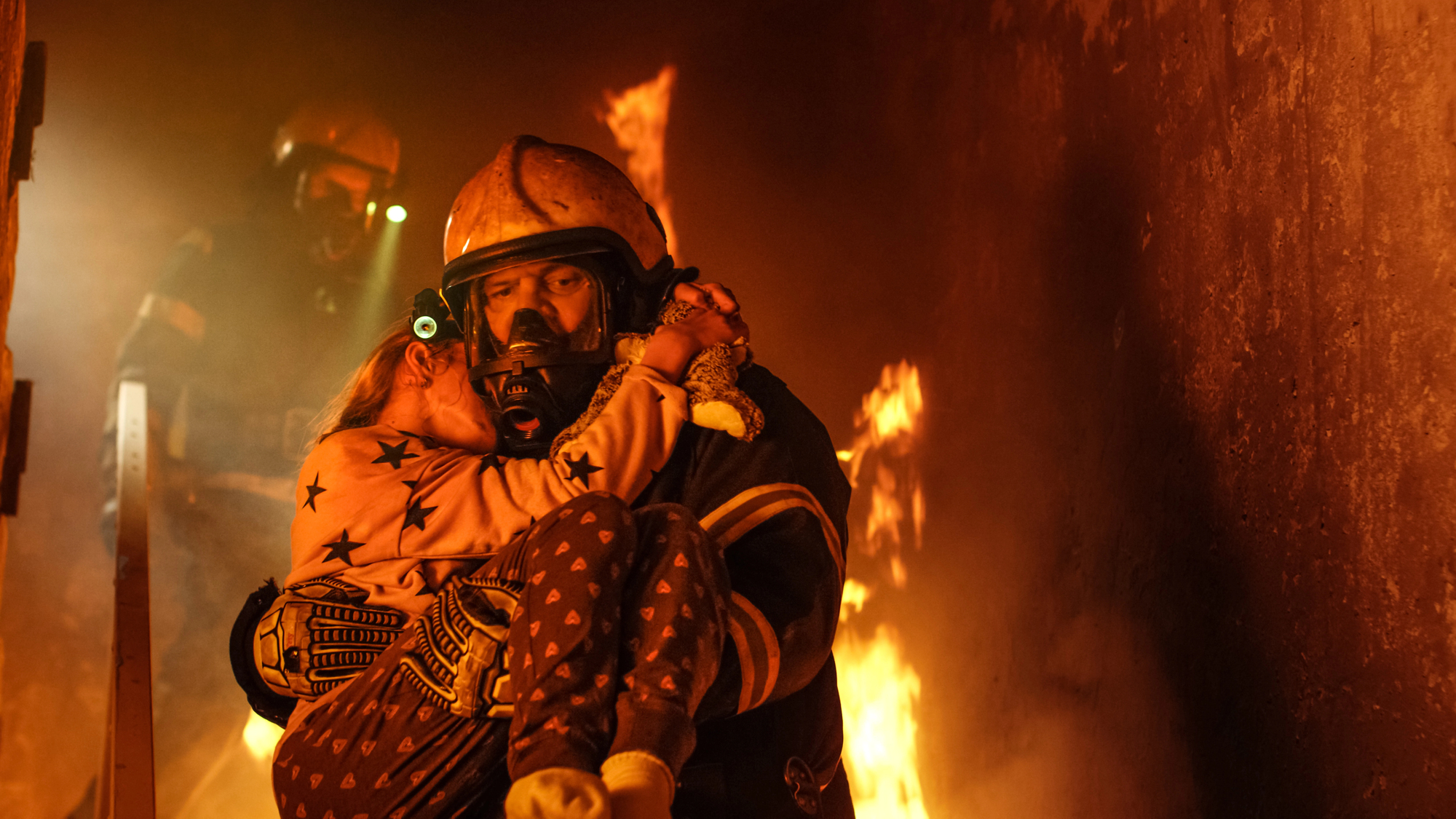 A firefighter wearing a helmet and protective gear carries a child through a burning building. Flames and smoke are visible around them. Another firefighter can be seen in the background on a ladder. The child clings to the firefighter, who is focused on the rescue.