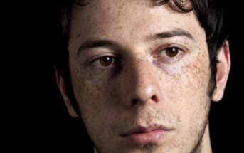 Close-up of a young man with fair skin, brown eyes, dark hair, and freckles. His expression is neutral, and he is looking slightly to the side. There is a black background behind him.