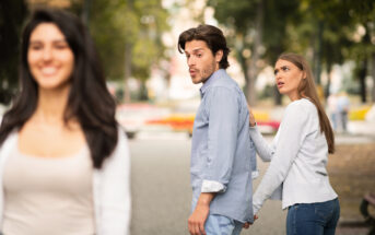 A man and a woman walk together while the man turns to look at another woman passing by, who is smiling and out of focus in the foreground. The woman with the man looks upset, holding onto his hand as they walk through a park.