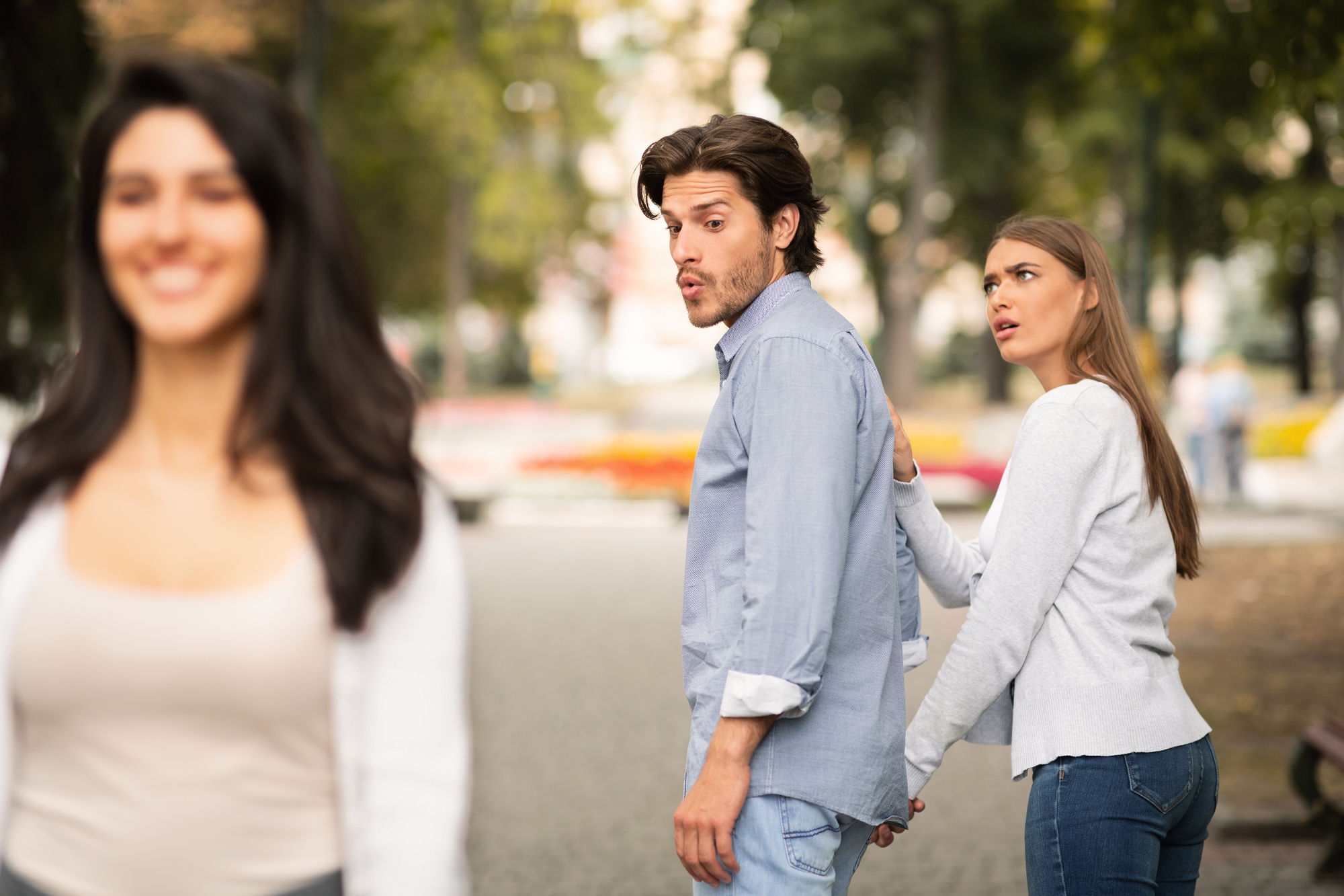 A man and a woman walk together while the man turns to look at another woman passing by, who is smiling and out of focus in the foreground. The woman with the man looks upset, holding onto his hand as they walk through a park.