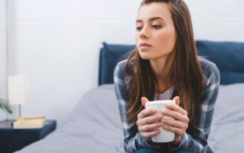 A young woman with long brown hair, wearing a plaid shirt, sits on a bed holding a white mug with both hands. She gazes thoughtfully to her left. The room has a minimalistic decor with a grey bedspread and a small bedside table with books and a lamp.