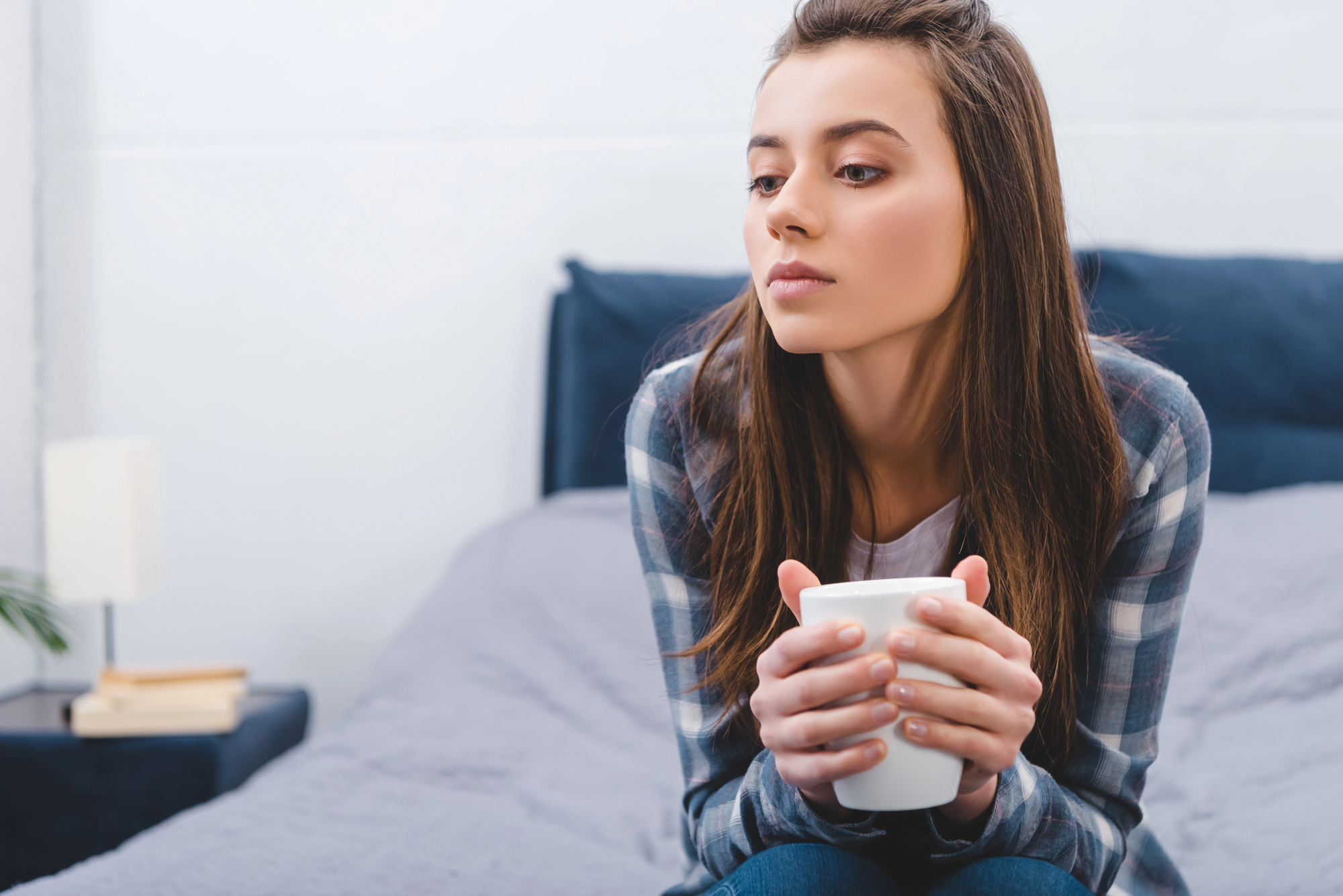 A young woman with long brown hair, wearing a plaid shirt, sits on a bed holding a white mug with both hands. She gazes thoughtfully to her left. The room has a minimalistic decor with a grey bedspread and a small bedside table with books and a lamp.