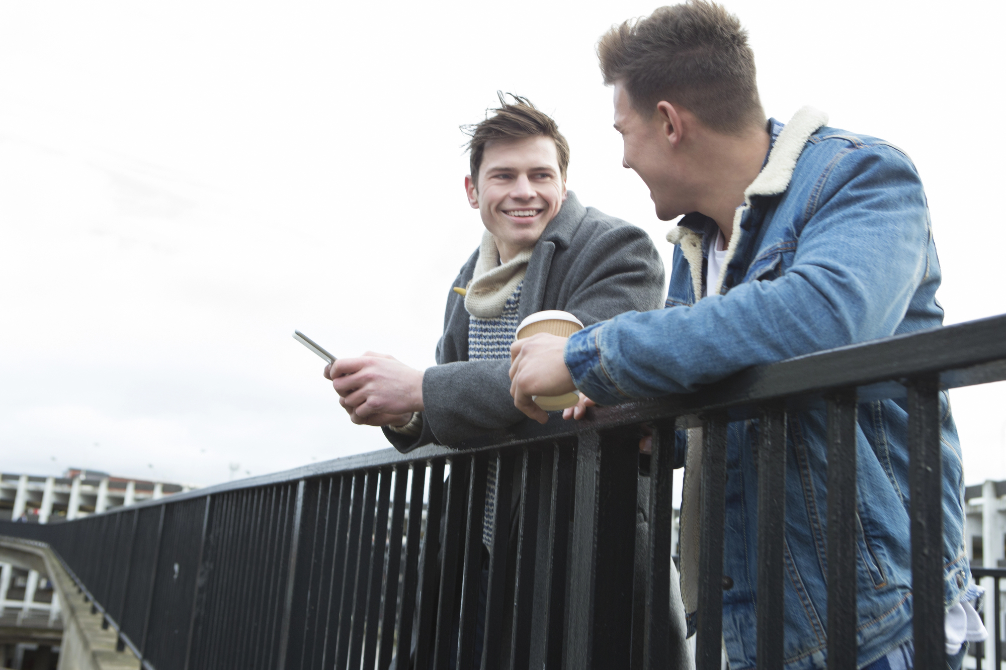 Two men are standing outdoors, leaning on a black railing. One man is holding a coffee cup while looking at the other, who holds a smartphone and smiles. Both are dressed casually in jackets with sherpa linings. A building and cloudy sky are visible in the background.