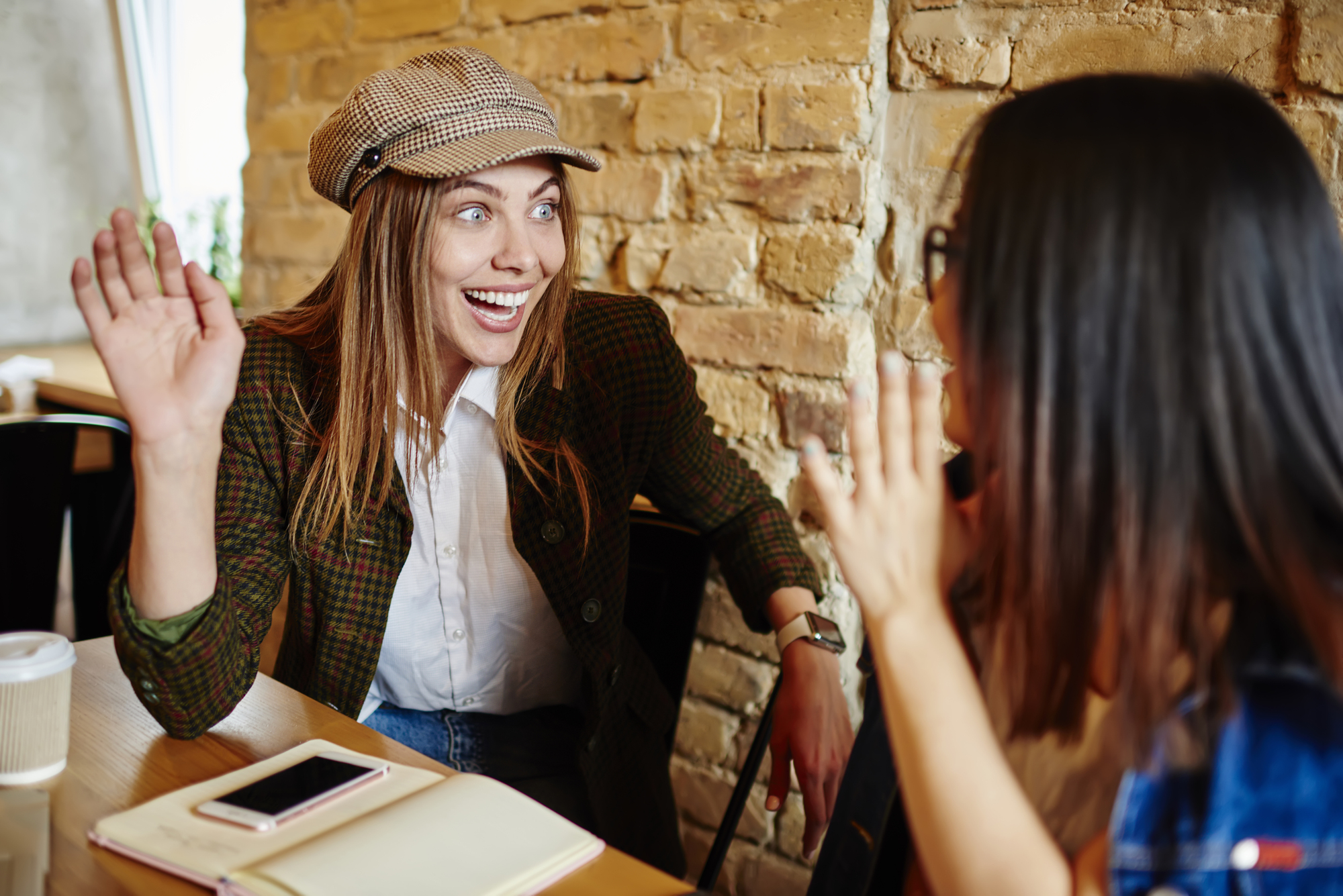 Two women seated at a wooden table in a café are enthusiastically greeting each other. One woman, wearing a brown plaid cap and jacket, has an expressive smile and raised hand. The other woman, wearing glasses, mirrors the gesture. A smartphone and notepad are on the table.