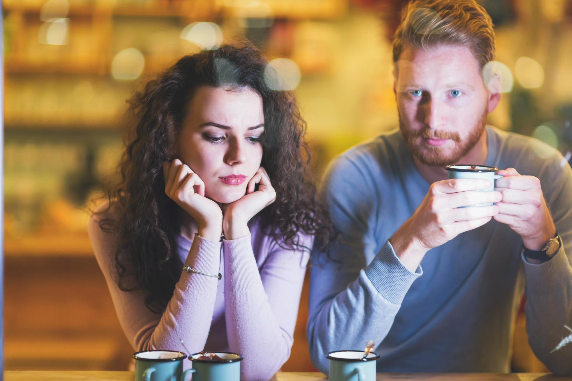 Two people are seated at a table in a cozy café. The woman on the left has curly hair and is resting her face on her hands, looking down and appearing thoughtful. The man on the right has a beard and is holding a cup while looking contemplative.