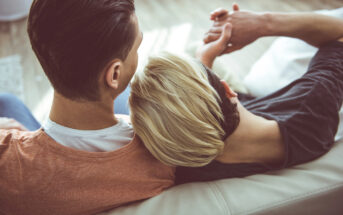 Two people are sitting close together on a couch. One leans their head on the other's shoulder. The person on the left wears an orange shirt, while the person on the right, with short blond hair, wears a dark gray shirt. Both hold hands, conveying a sense of comfort and intimacy.