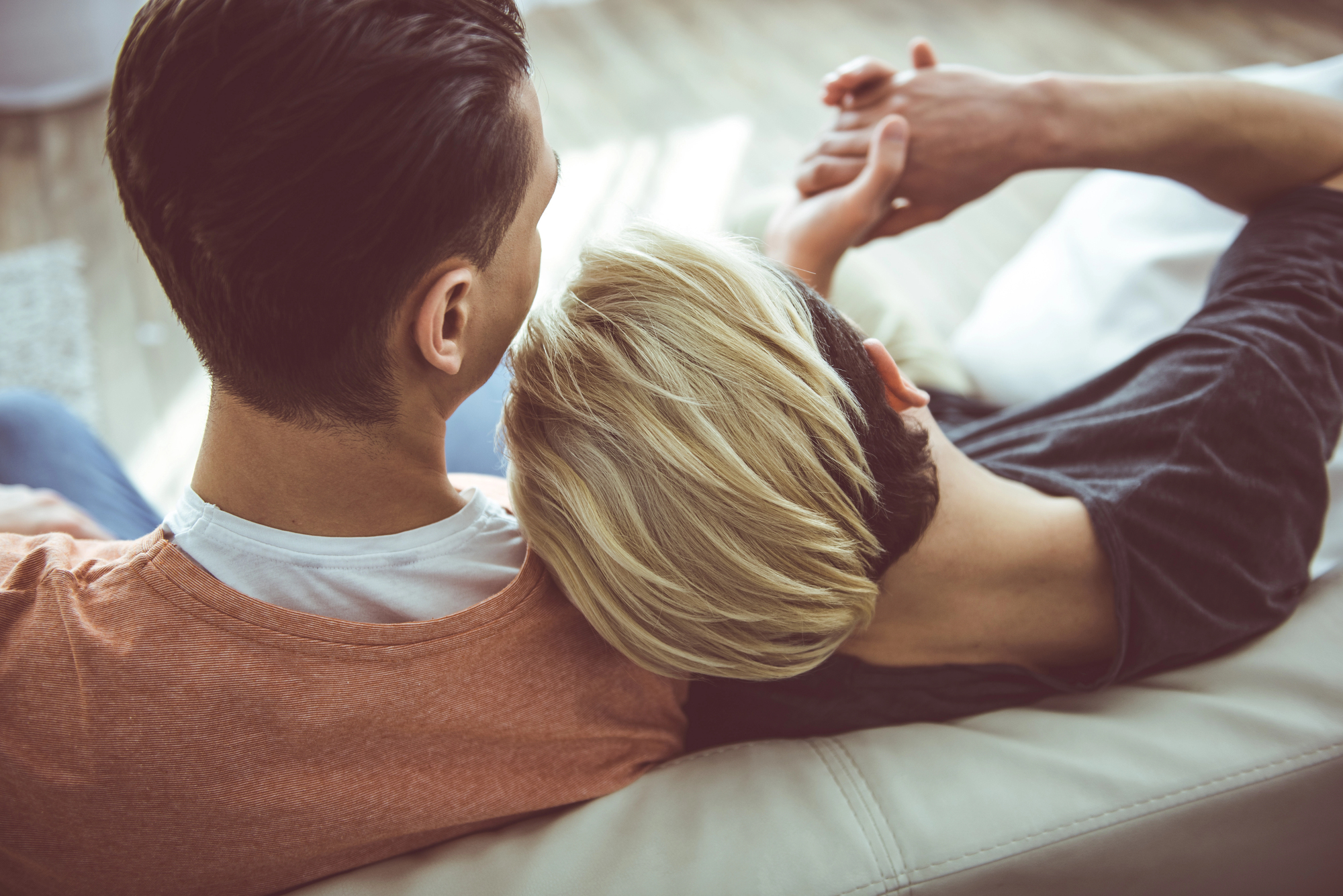 Two people are sitting close together on a couch. One leans their head on the other's shoulder. The person on the left wears an orange shirt, while the person on the right, with short blond hair, wears a dark gray shirt. Both hold hands, conveying a sense of comfort and intimacy.