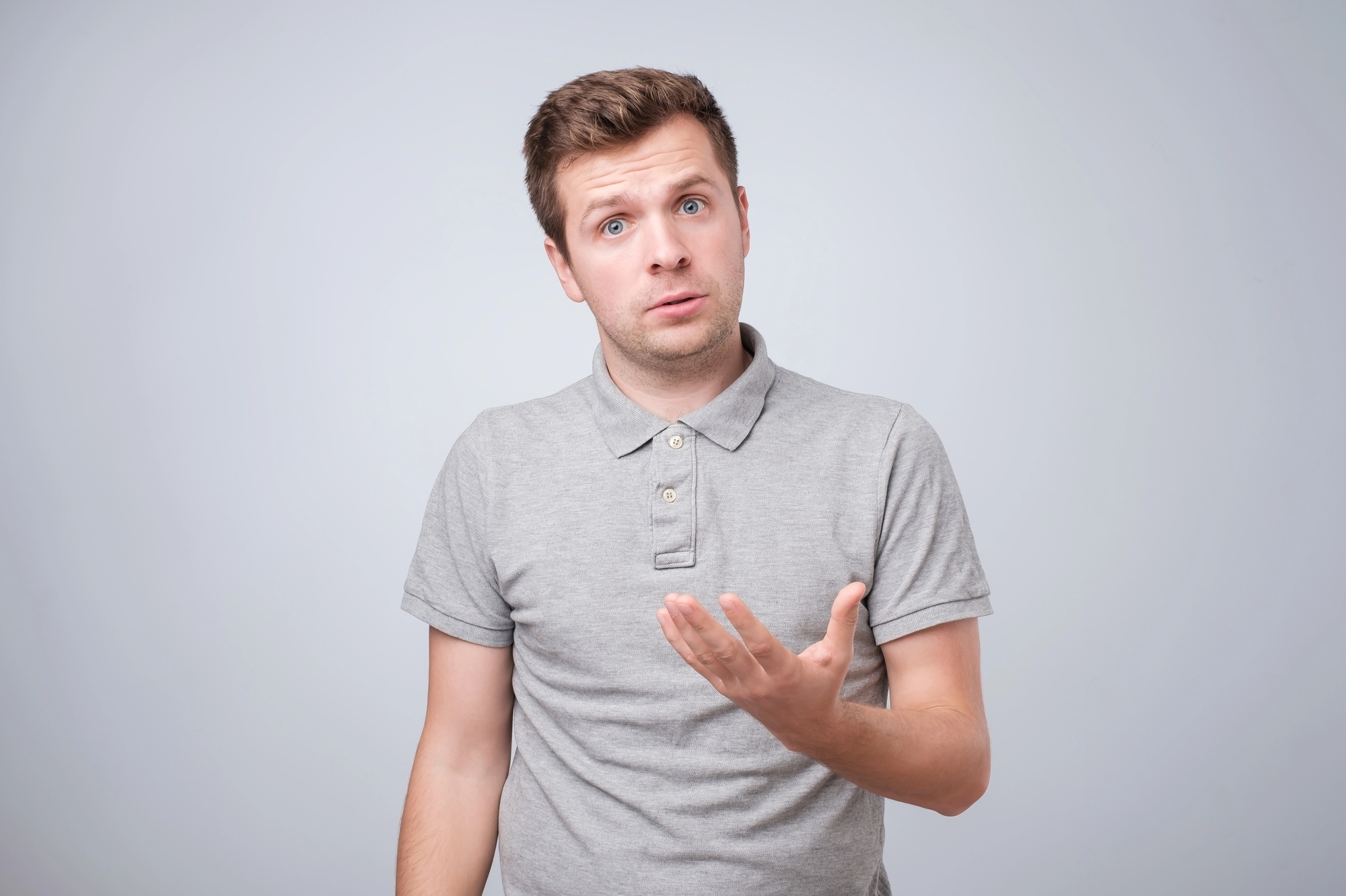 A man in a gray polo shirt stands against a plain gray background. He has a perplexed expression, with one eyebrow slightly raised and his mouth slightly open. His right hand is raised in an open gesture, as if questioning or explaining something.