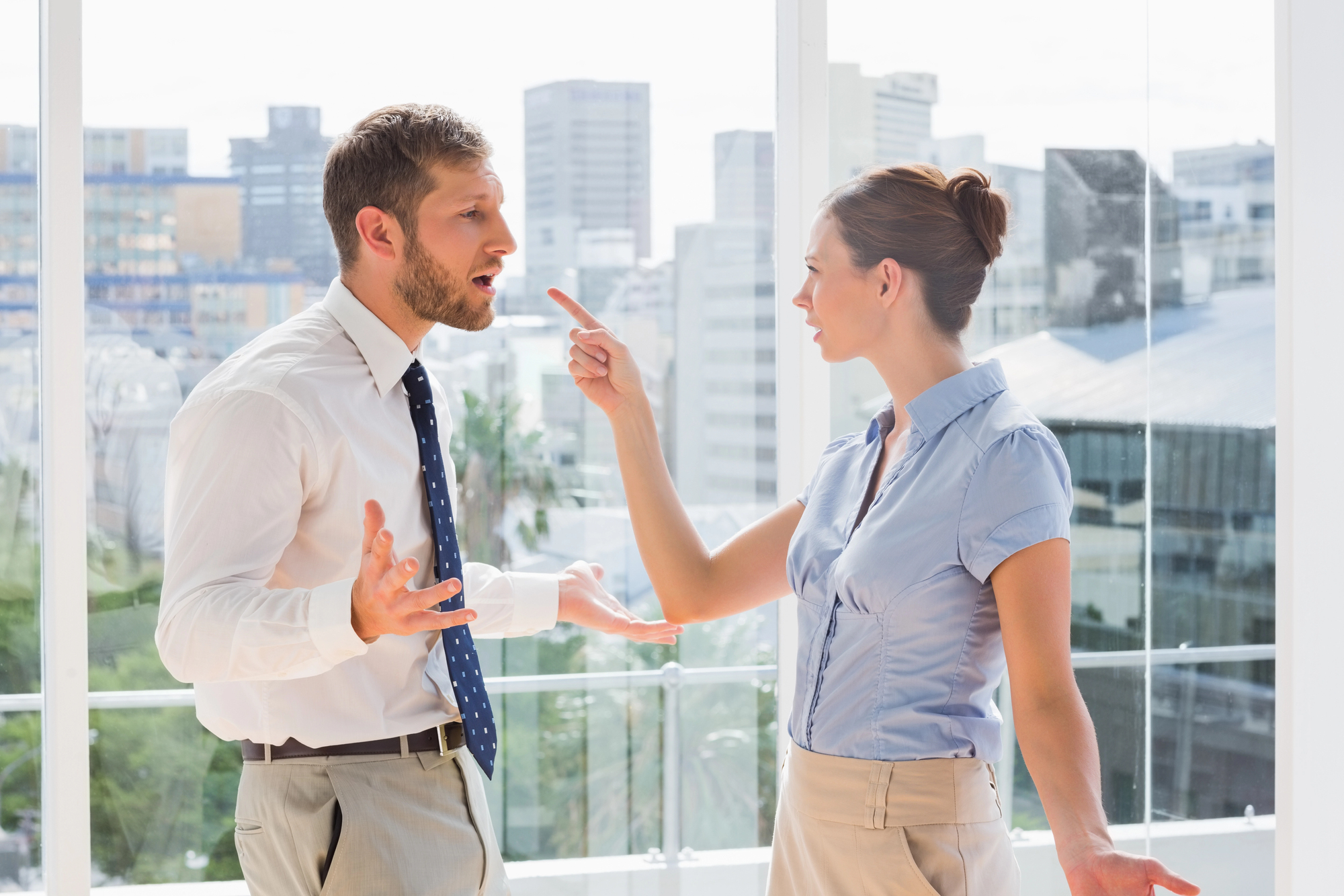 A man and a woman are having an intense conversation in a bright office with large windows overlooking a cityscape. The man looks surprised with his hands up, while the woman is pointing her finger at him, appearing to be frustrated. Both are dressed in business attire.