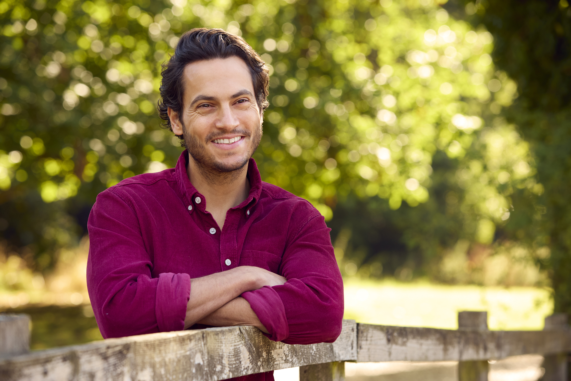 A man wearing a red shirt is leaning against a wooden fence with his arms crossed. He is smiling and standing in an outdoor setting with a green, leafy background, illuminated by sunlight.