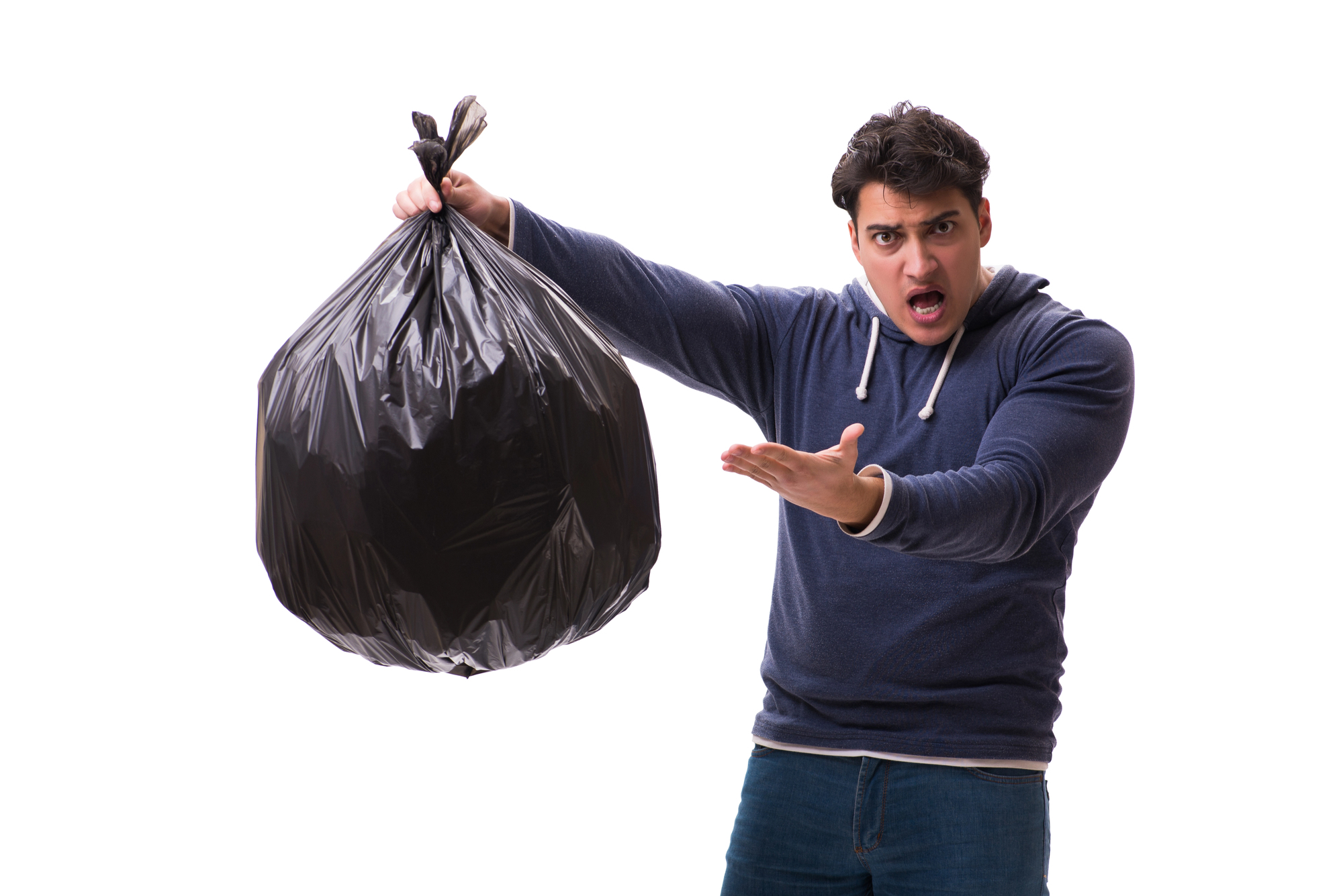 A man with a surprised expression holds up a large black garbage bag in his right hand and points to it with his left hand. He is wearing a blue hoodie and jeans and is standing against a white background.