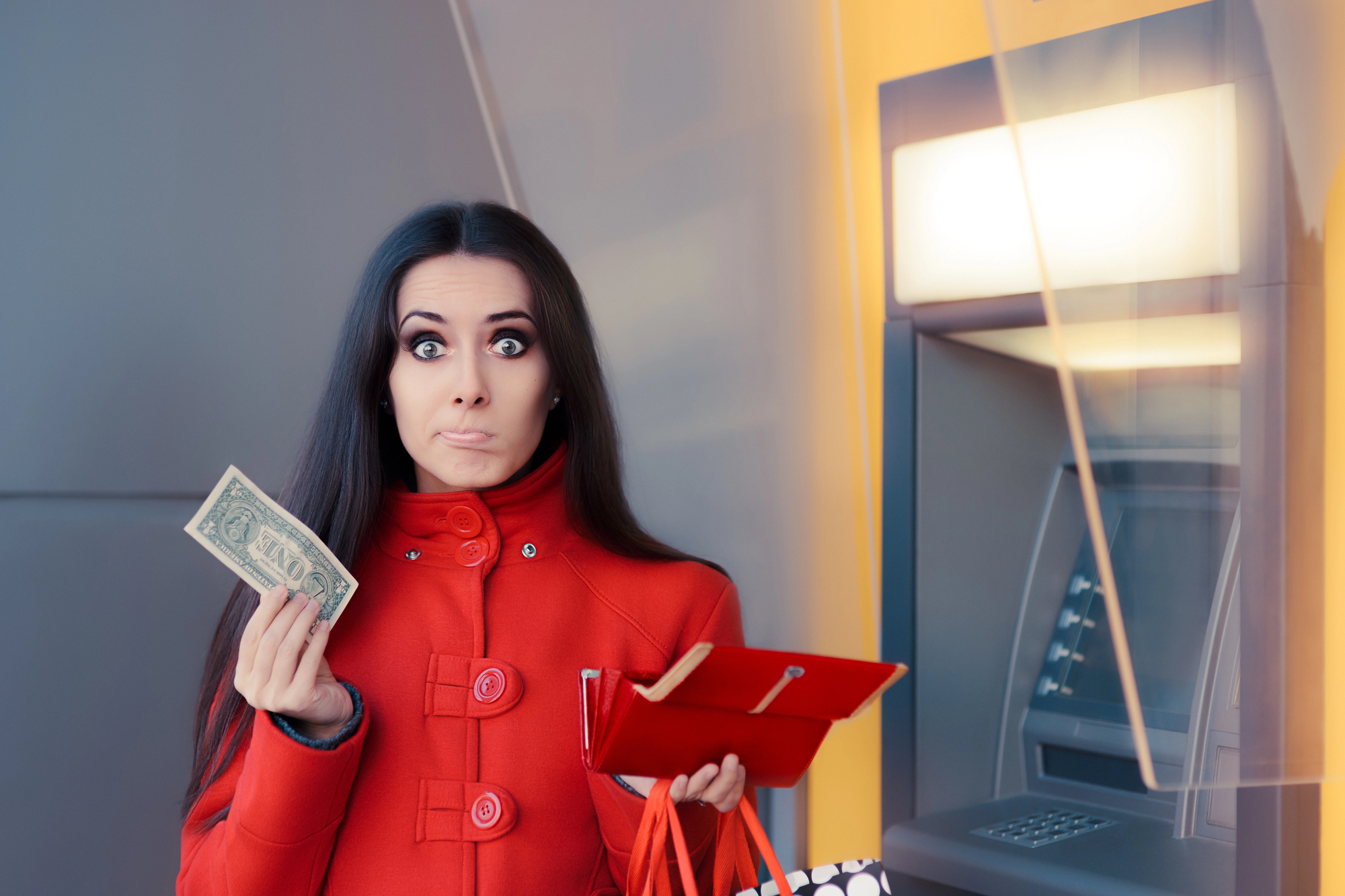 A woman in a red coat with an anxious expression holds a single dollar bill in one hand and an open wallet in the other. She stands next to an ATM, and shopping bags hang from her arm.