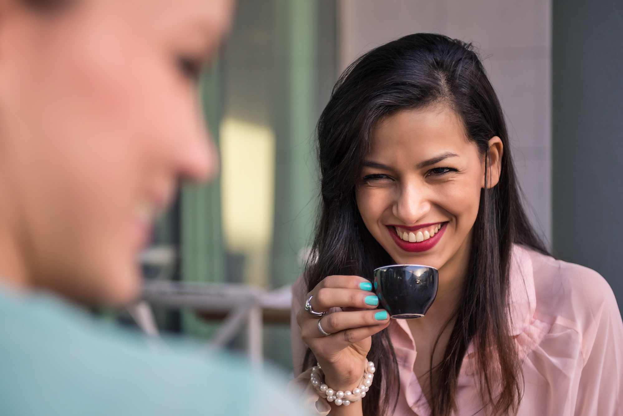 A woman with long dark hair, wearing a light pink blouse and bracelets, smiles warmly while holding a small espresso cup. Another person is blurred in the foreground. The setting appears casual and pleasant.