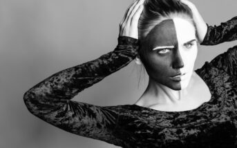 A monochrome image of a person with face paint, divided into black and white halves down the middle. They are wearing a velvet-textured top and pose with both hands on their head, one eyebrow raised, creating a dramatic visual effect.