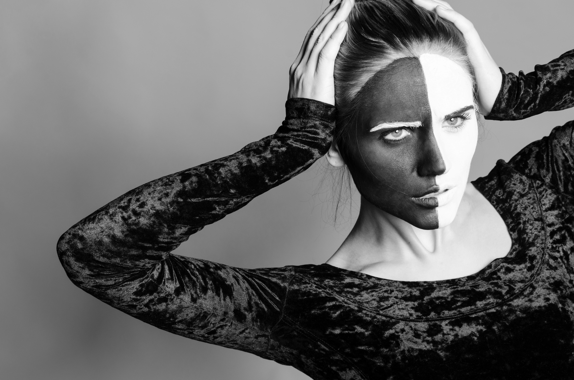 A monochrome image of a person with face paint, divided into black and white halves down the middle. They are wearing a velvet-textured top and pose with both hands on their head, one eyebrow raised, creating a dramatic visual effect.