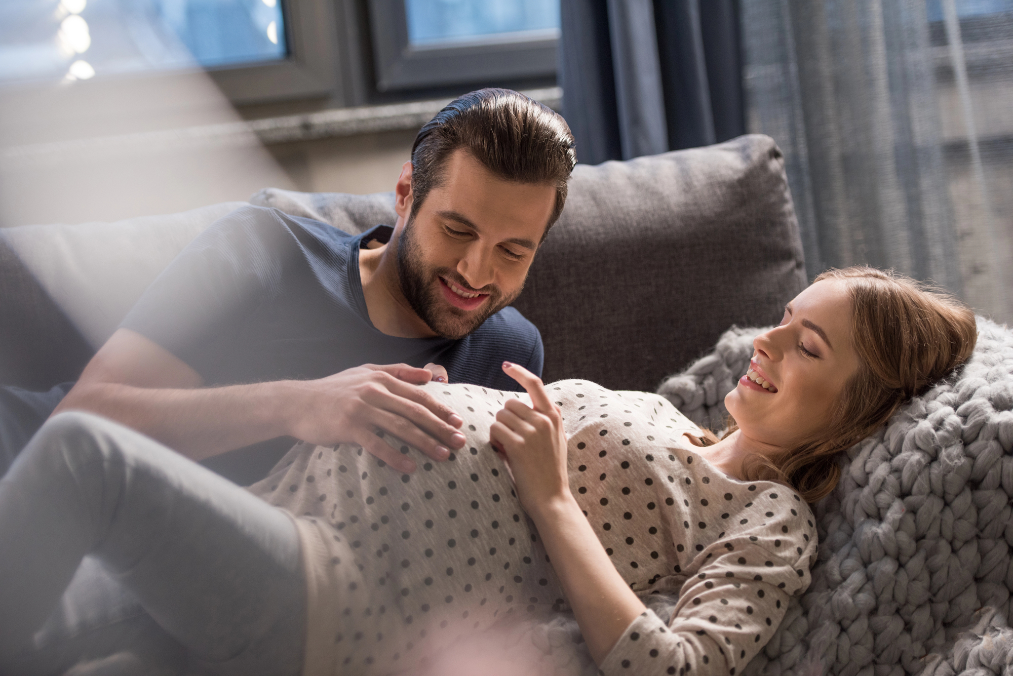 A man and a pregnant woman are lying on a couch, smiling and looking at each other. The man gently places his hand on the woman's baby bump, while the woman points at her belly. They are in a cozy indoor setting with soft lighting.