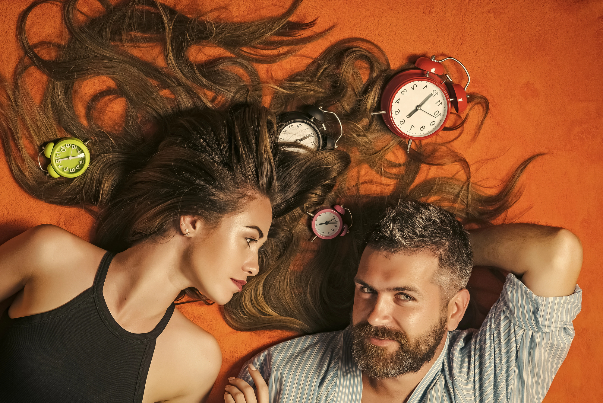 A woman and a man lie on an orange surface. The woman's long hair is spread out around several old-fashioned alarm clocks of various colors. She has a thoughtful expression while the man, with a beard, rests his head on his hand, looking content.