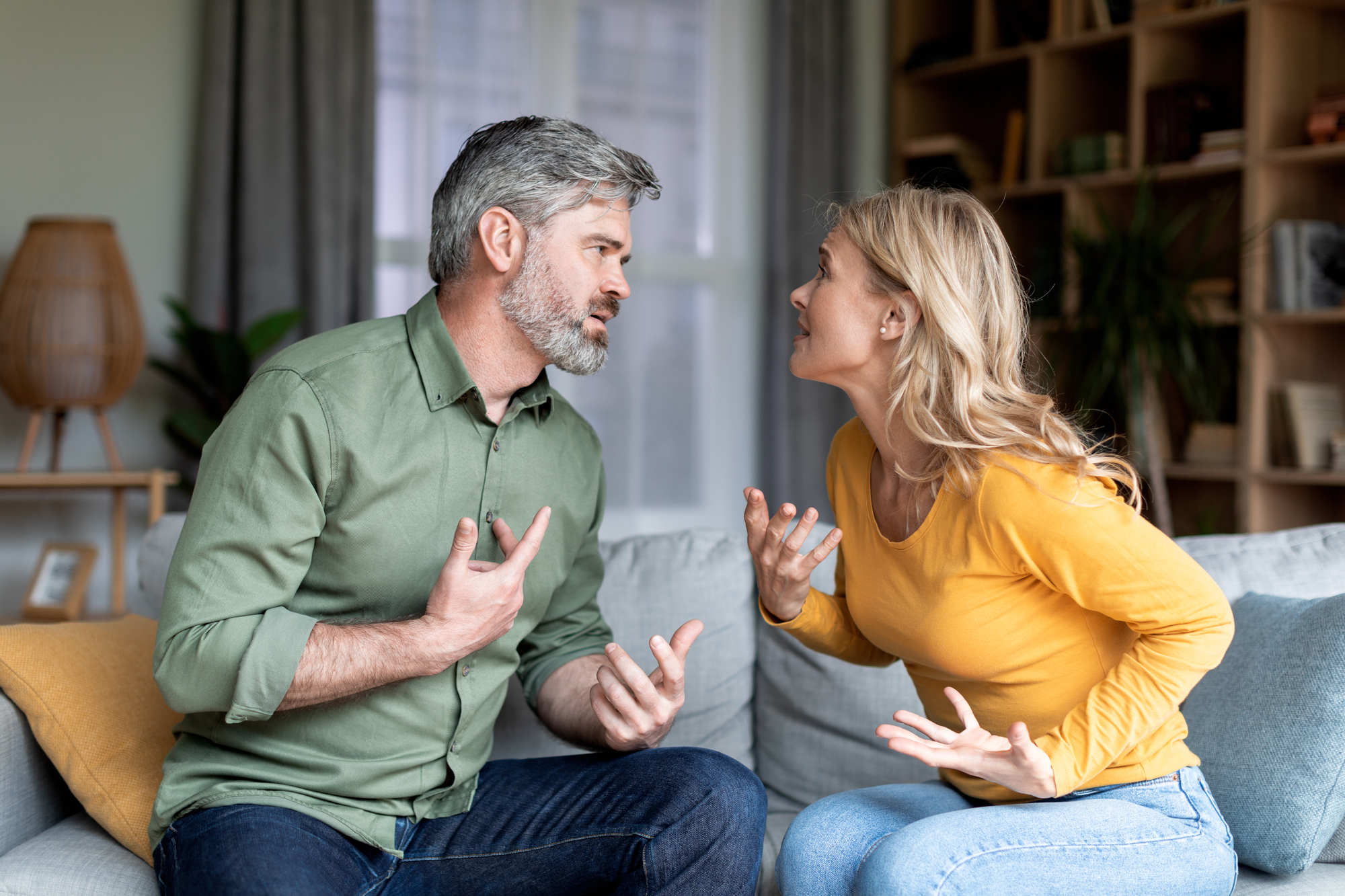 A middle-aged man with gray hair and beard wearing a green shirt, and a blonde middle-aged woman in a yellow shirt, are sitting on a couch inside a cozy living room. They are arguing passionately, gesturing with their hands, and looking at each other intensely.