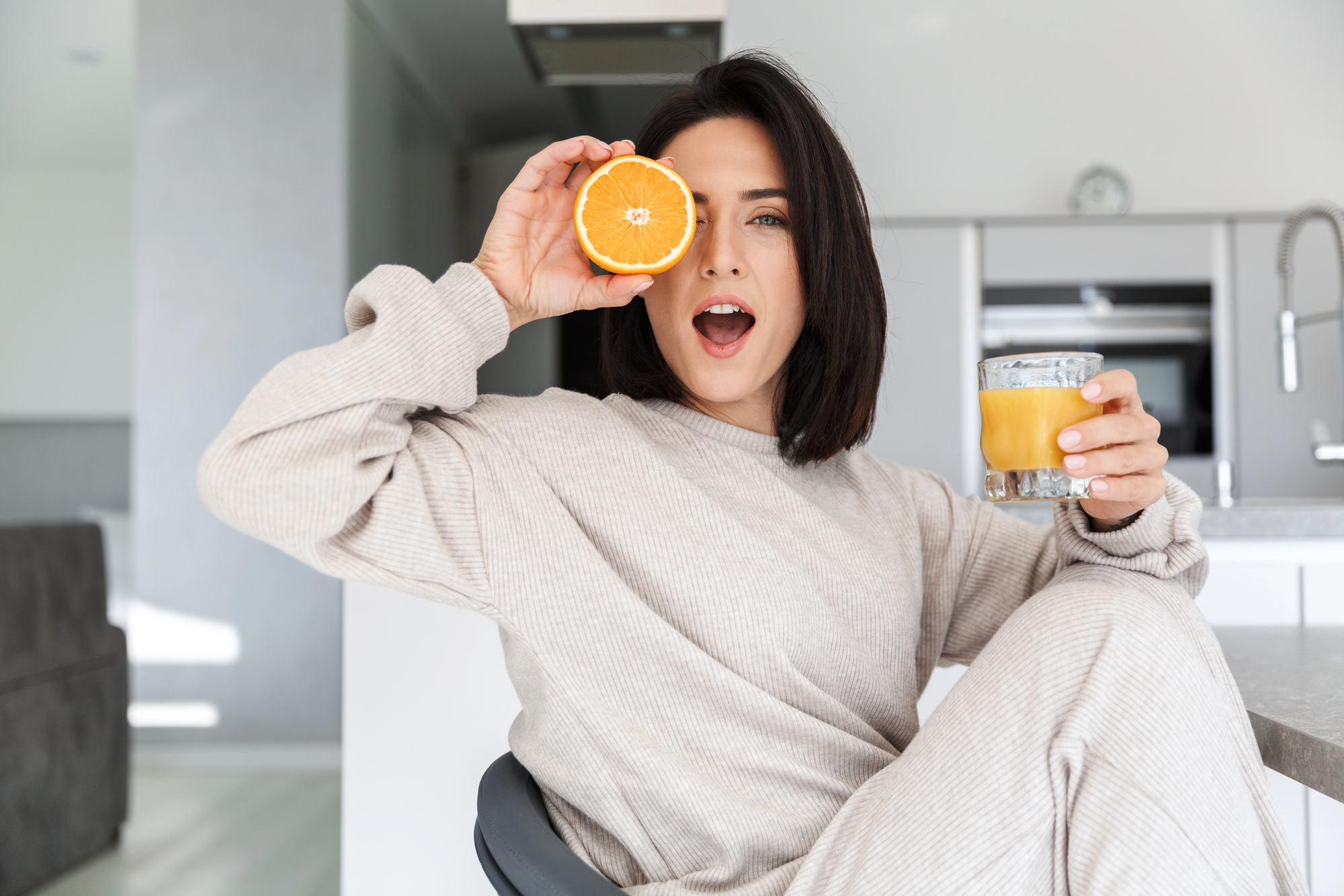 A woman with medium-length dark hair, wearing a light beige sweater and pants, is sitting on a chair in a modern kitchen. She is holding an orange slice in front of one eye and a glass of orange juice in her other hand, looking surprised and cheerful.