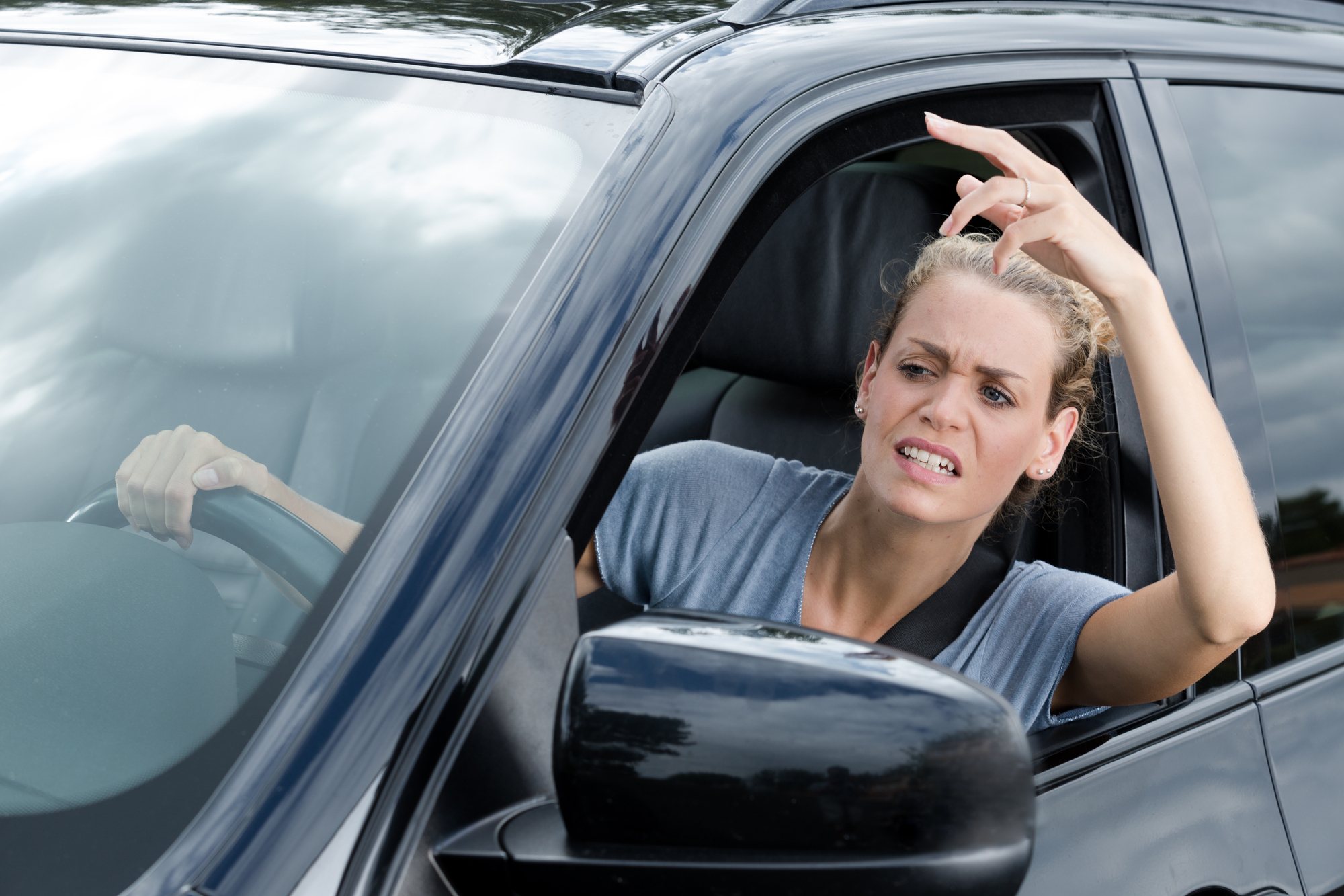 A woman driving a black car looks frustrated and is gesturing with her hand out the window. She has a tense facial expression and appears to be upset about something on the road. The background shows an overcast sky.