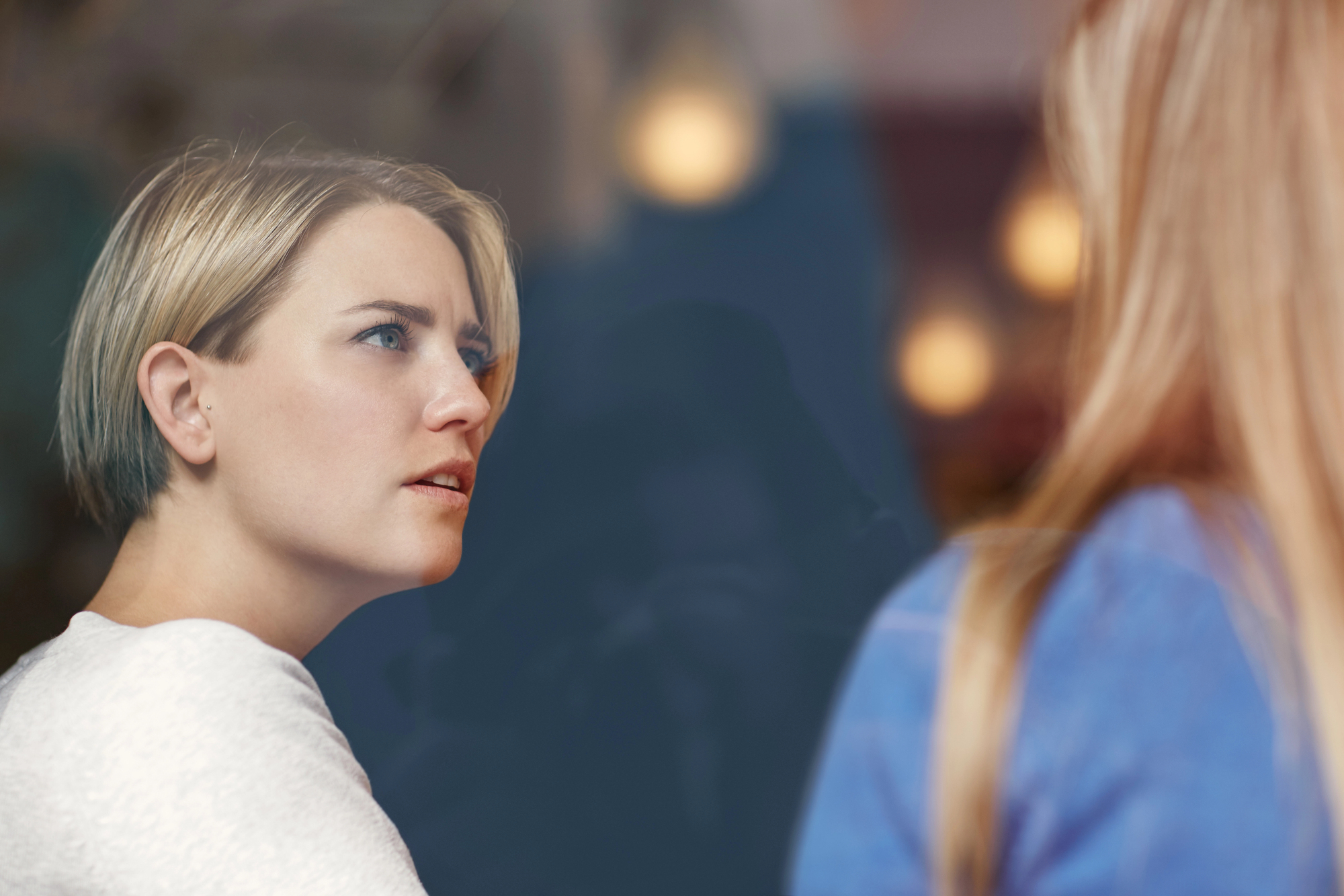 A woman with short blond hair, wearing a white sweater, is looking thoughtfully to her right. She is seen through a reflective glass, and another person with long blond hair, partially out of focus, is in the foreground facing away.