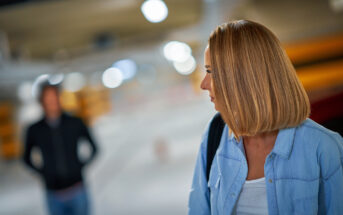 A woman with shoulder-length blonde hair wearing a blue shirt looks over her shoulder in a parking garage. A hooded figure stands in the background, slightly blurred. The scene appears tense or suspenseful.