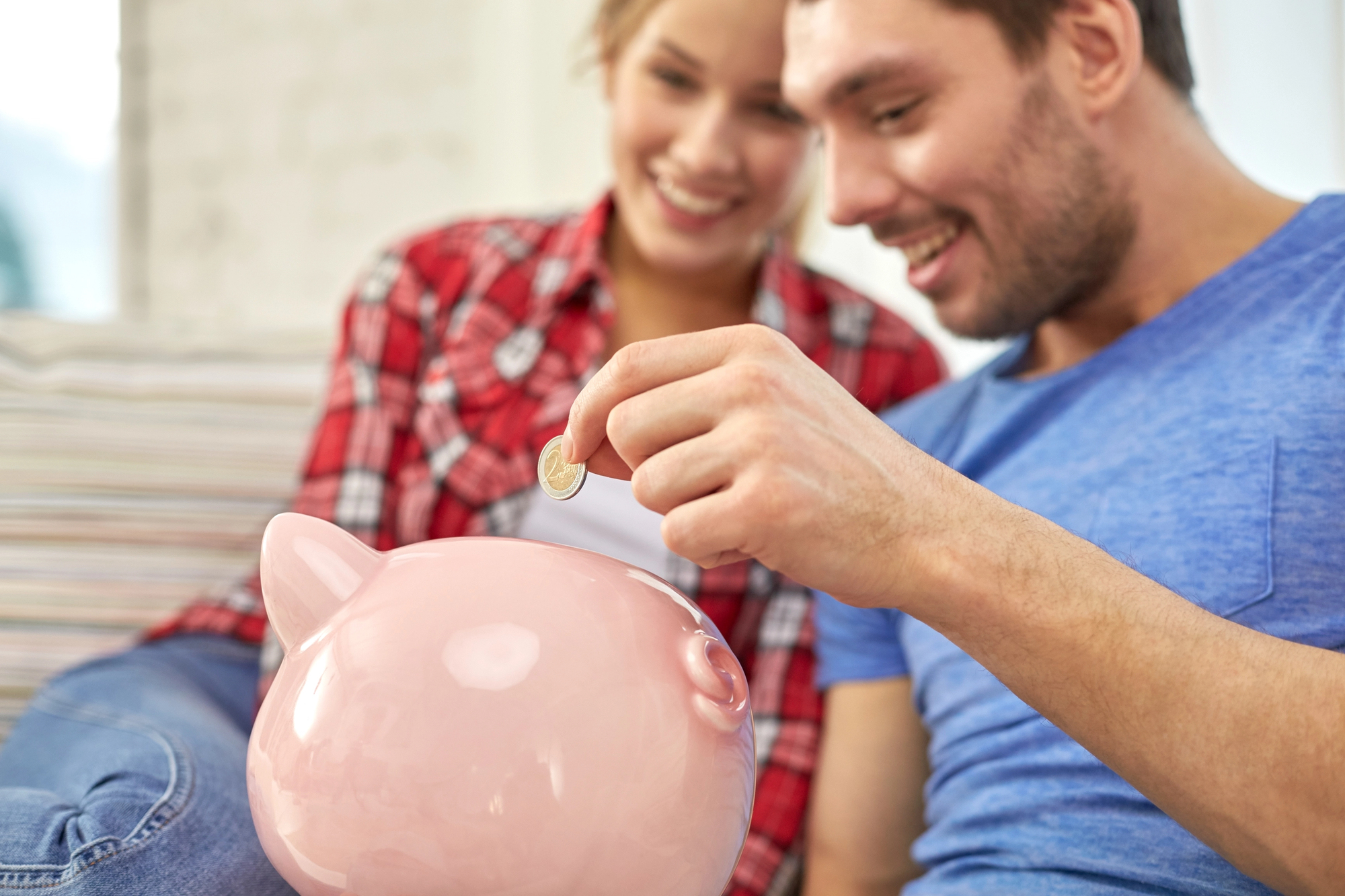 A smiling couple sits on a couch, both looking at a large pink piggy bank. The man, wearing a blue shirt, is about to drop a coin into the piggy bank. The woman, in a red plaid shirt, watches closely. They appear happy and engaged in saving money.