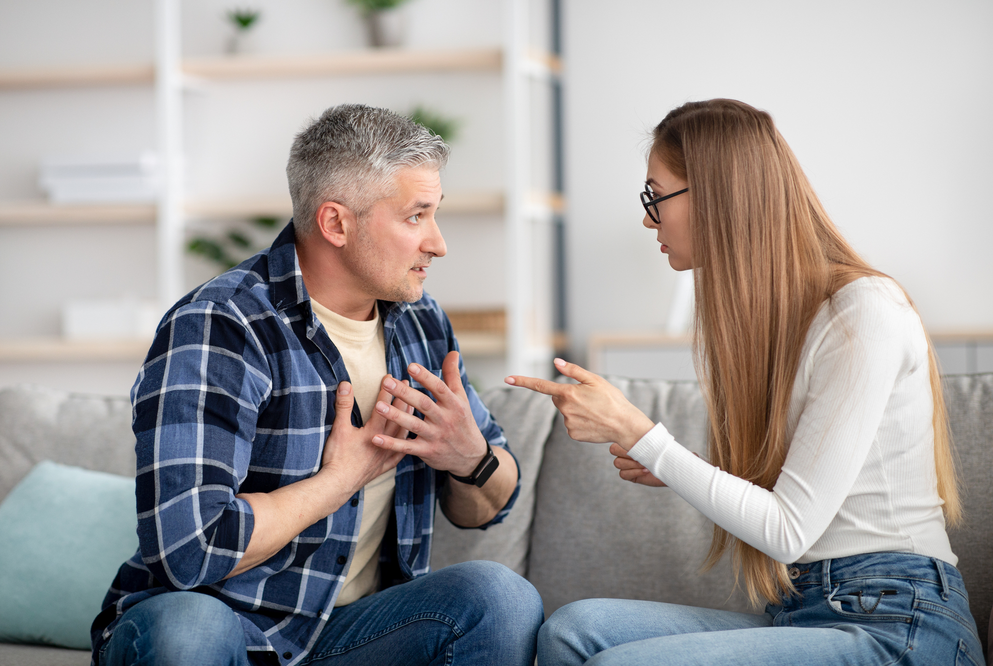 A man with gray hair and wearing a blue plaid shirt and jeans, sits on a couch with a concerned expression. A woman with long blond hair, a white sweater, and jeans, wearing glasses, sits beside him, pointing her finger and looking frustrated. Shelves are visible in the background.