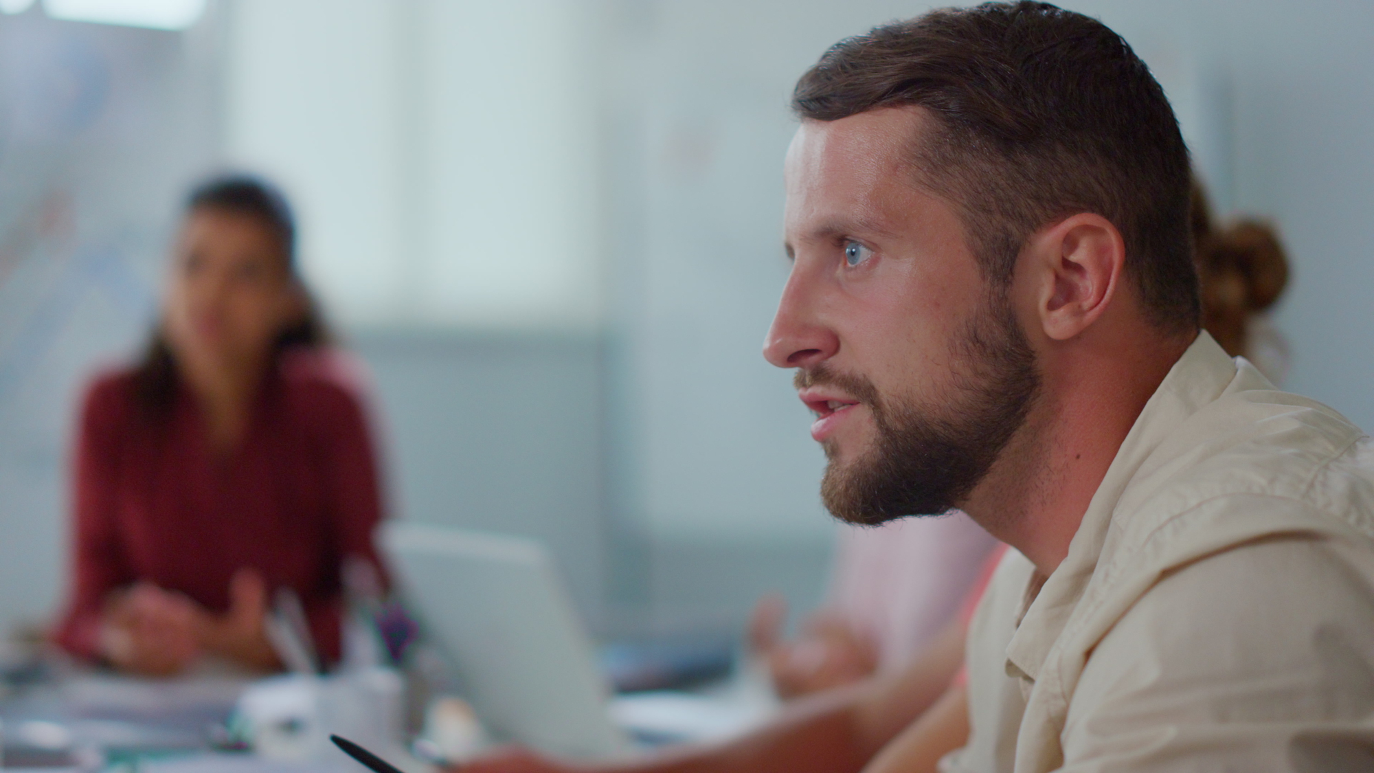 A focused man with short brown hair and a beard is seated at a desk in an office, holding a pen. In the background, there are blurred people and office equipment. The setting appears to be a meeting or a work discussion.